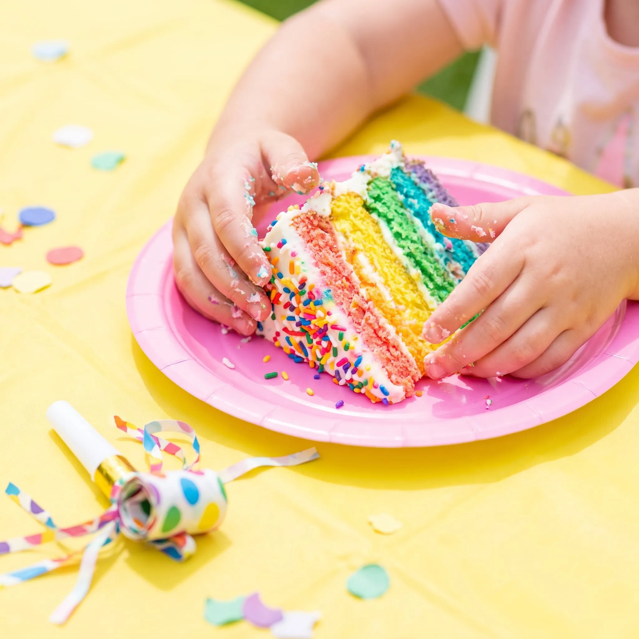 Small hands holding a slice of rainbow layer cake with sprinkles on a pink paper plate