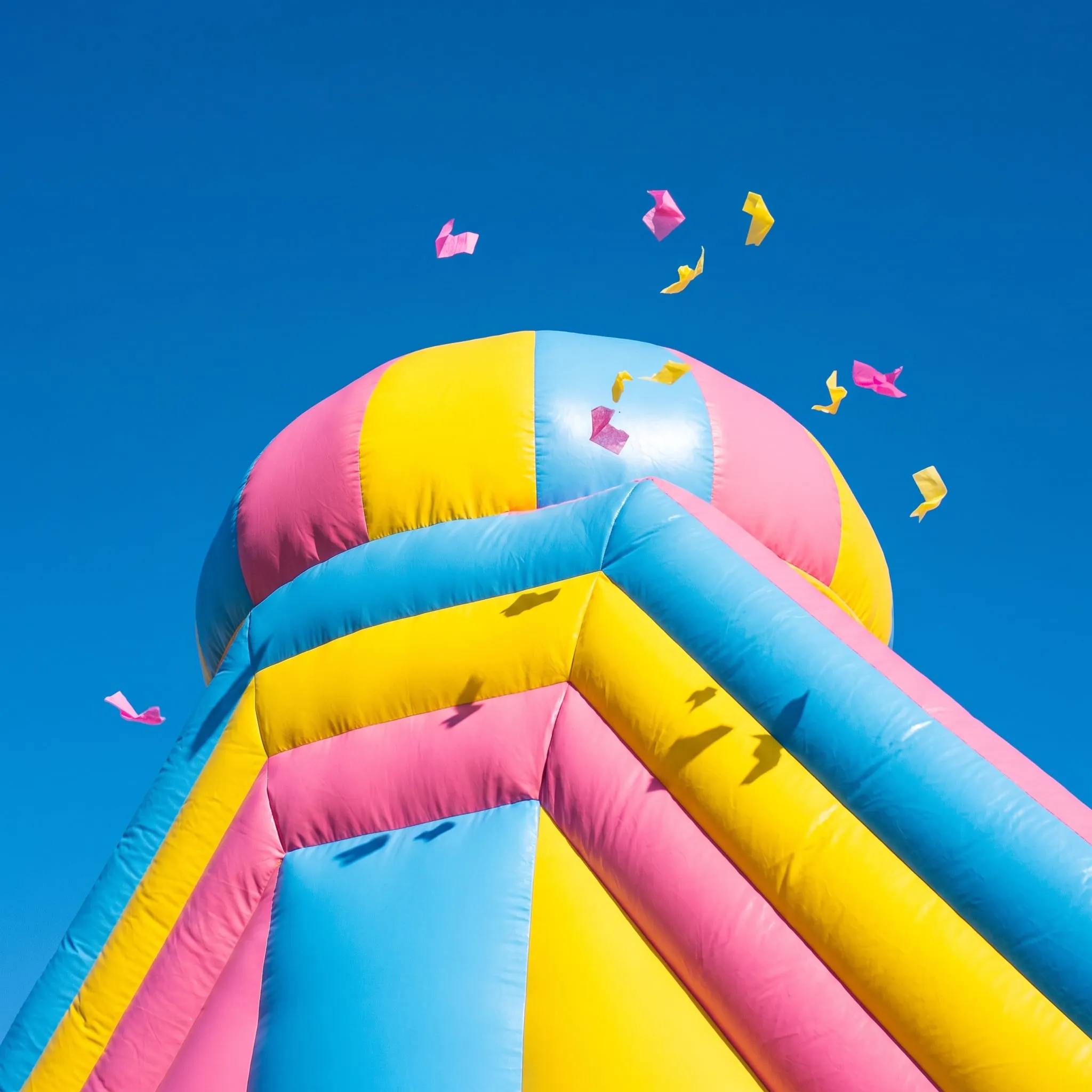 The top of a pink, yellow and blue bouncy castle against a blue sky with confetti in the air