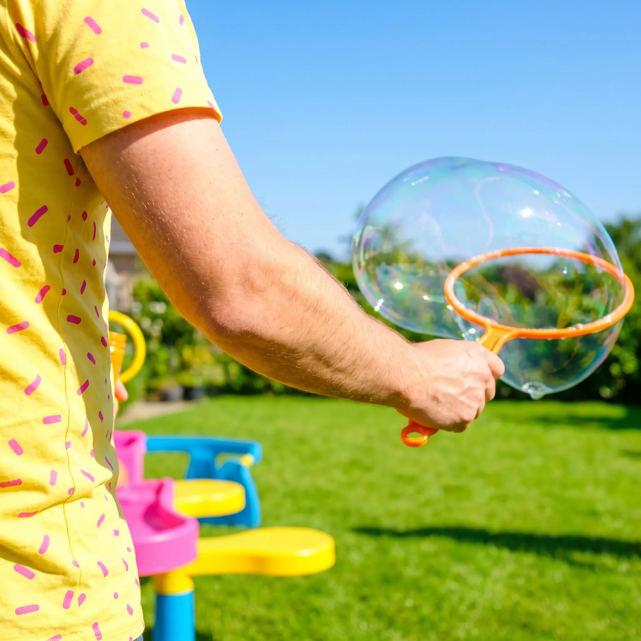 A host's arm in a yellow confetti-print shirt making a giant bubble over a backyard lawn