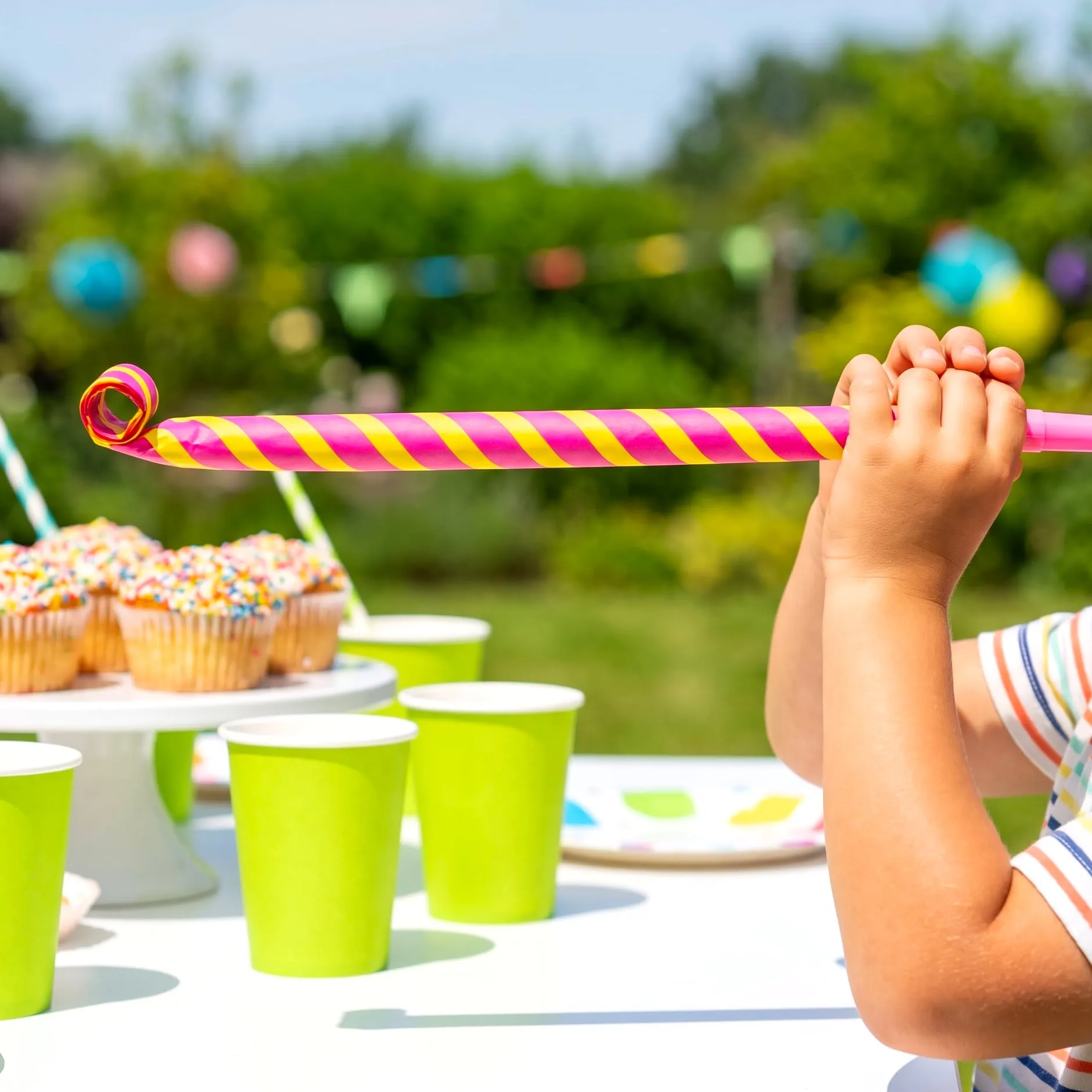Small hands holding a striped pink and yellow party blower over a table of sprinkle cupcakes and green cups