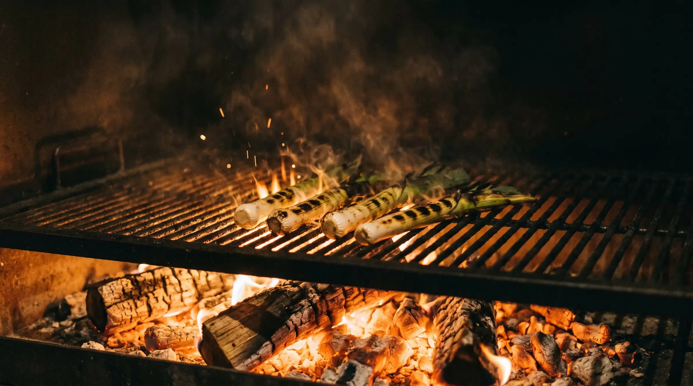 Young leeks charring on a steel grill over glowing hardwood coals, sparks and smoke rising.