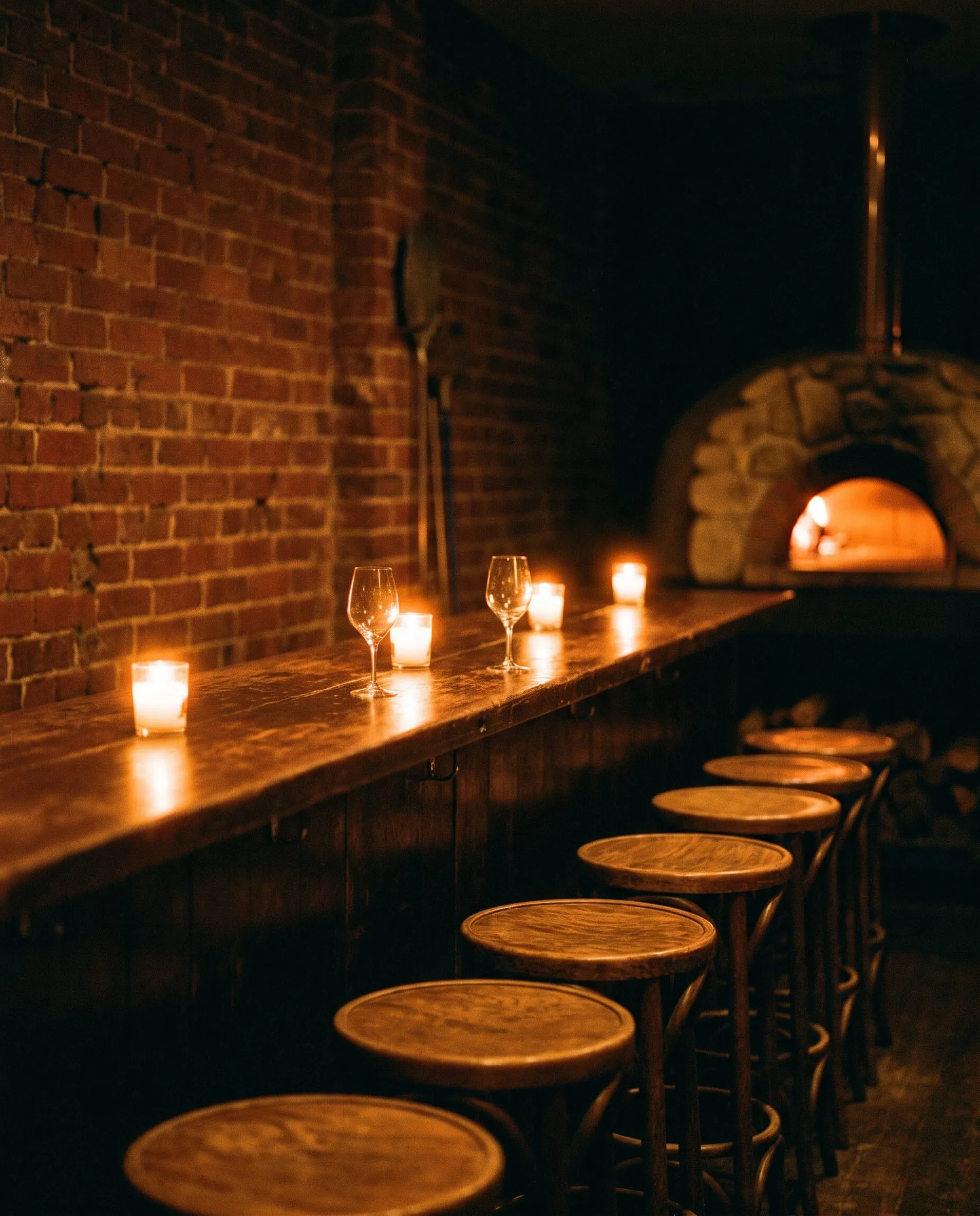 A row of empty timber bar stools along a candlelit bar, a wood oven glowing at the far end of the room.