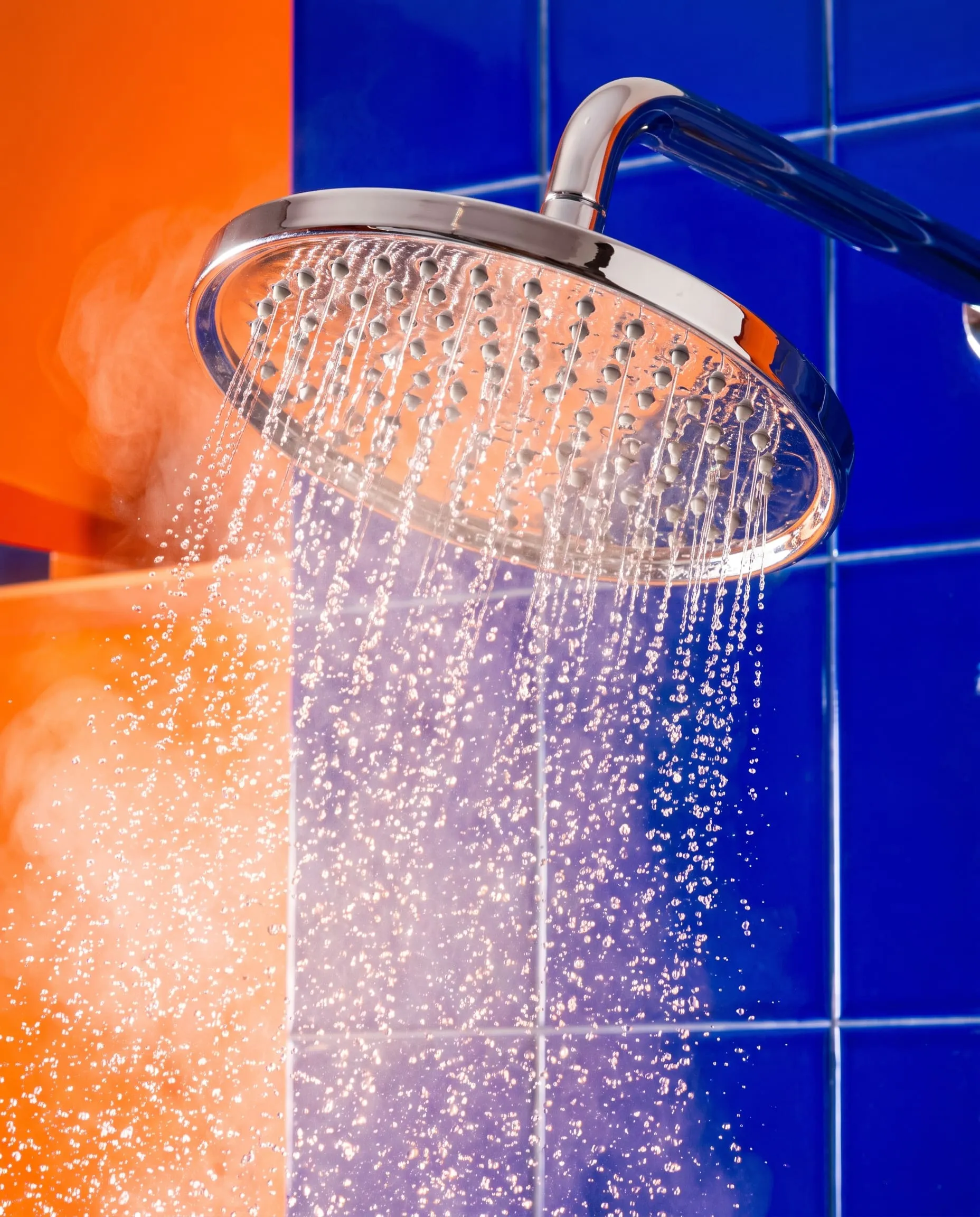 A chrome rain shower head spraying steaming water against blue tiles