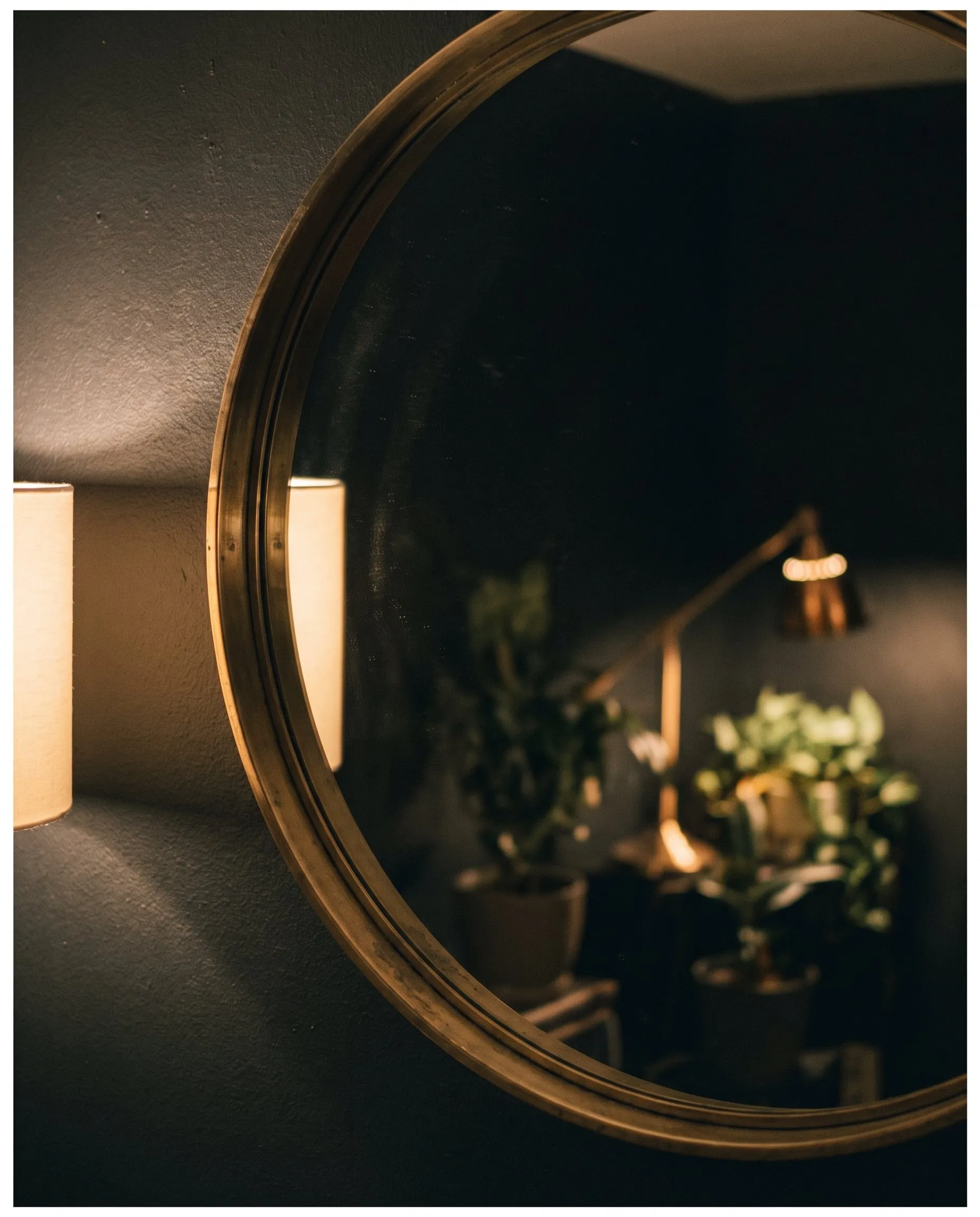 A round brass-framed mirror on a charcoal wall reflecting plants and warm light