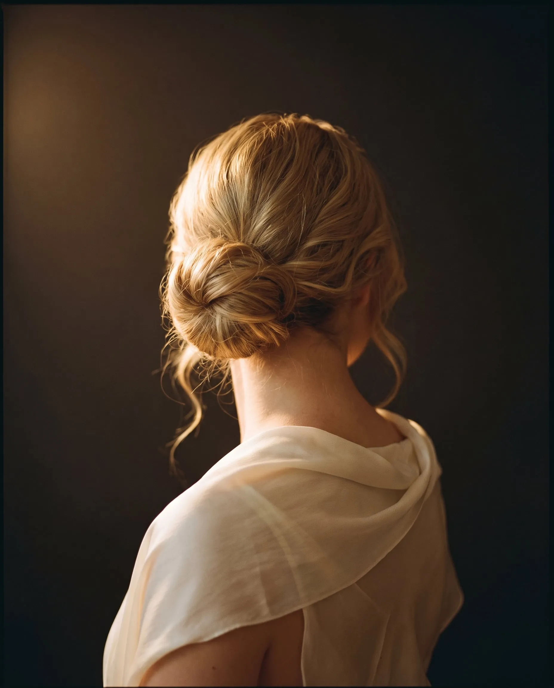 A soft low bun in golden blonde with loose tendrils at the nape, ivory silk at the shoulders