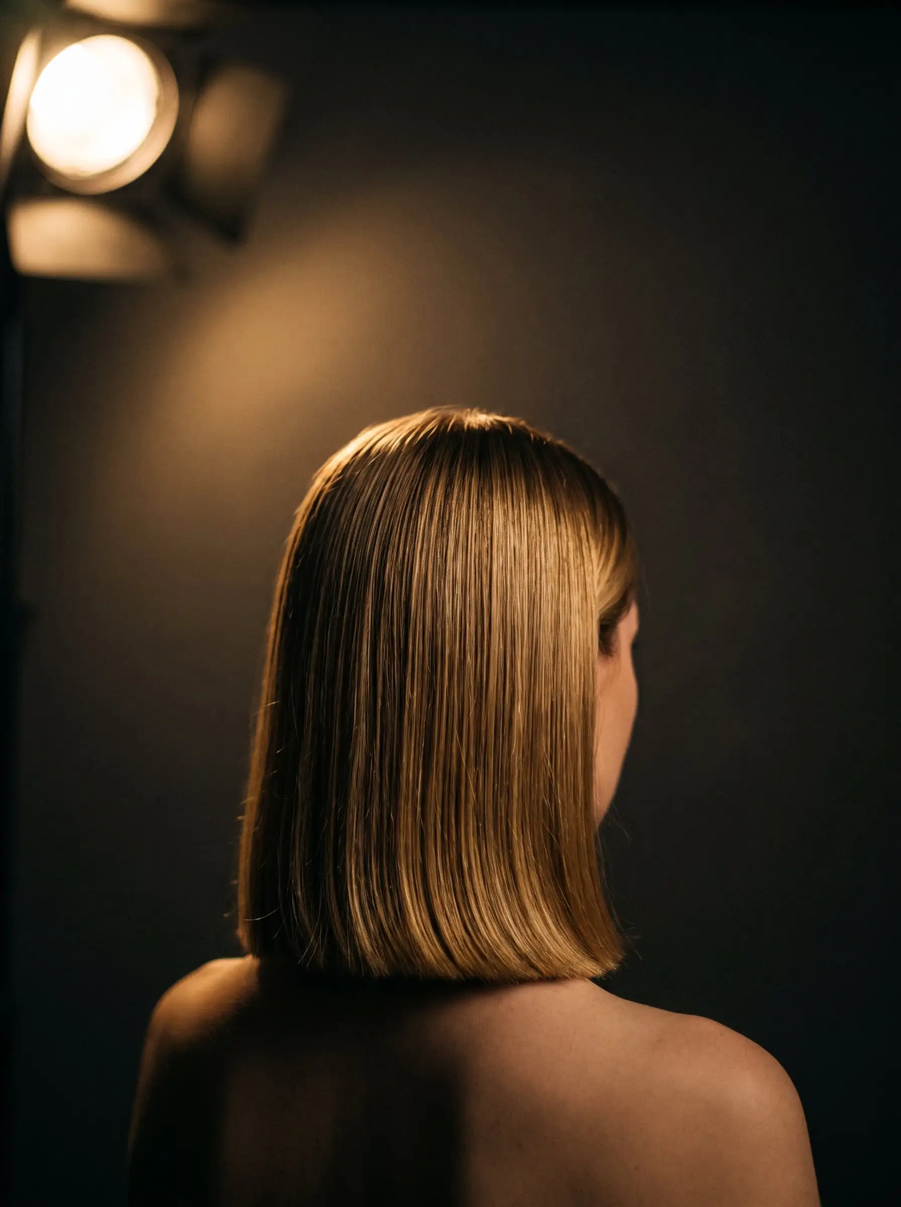 A sleek, blunt shoulder-length cut in golden dark blonde, seen from behind under a warm studio light