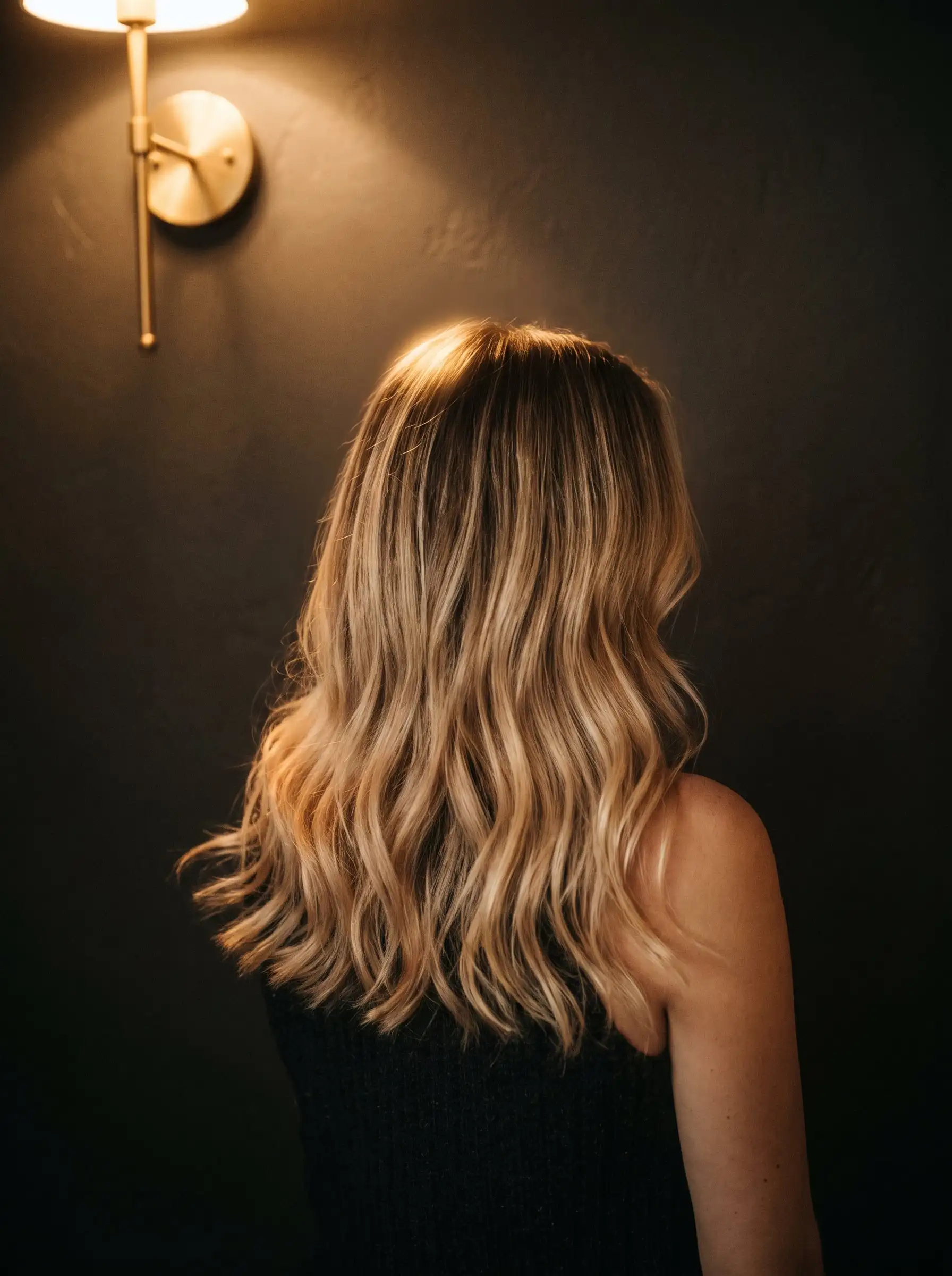 Long lived-in blonde hair with soft waves, seen from behind, catching the warm light of a brass wall lamp against a charcoal wall