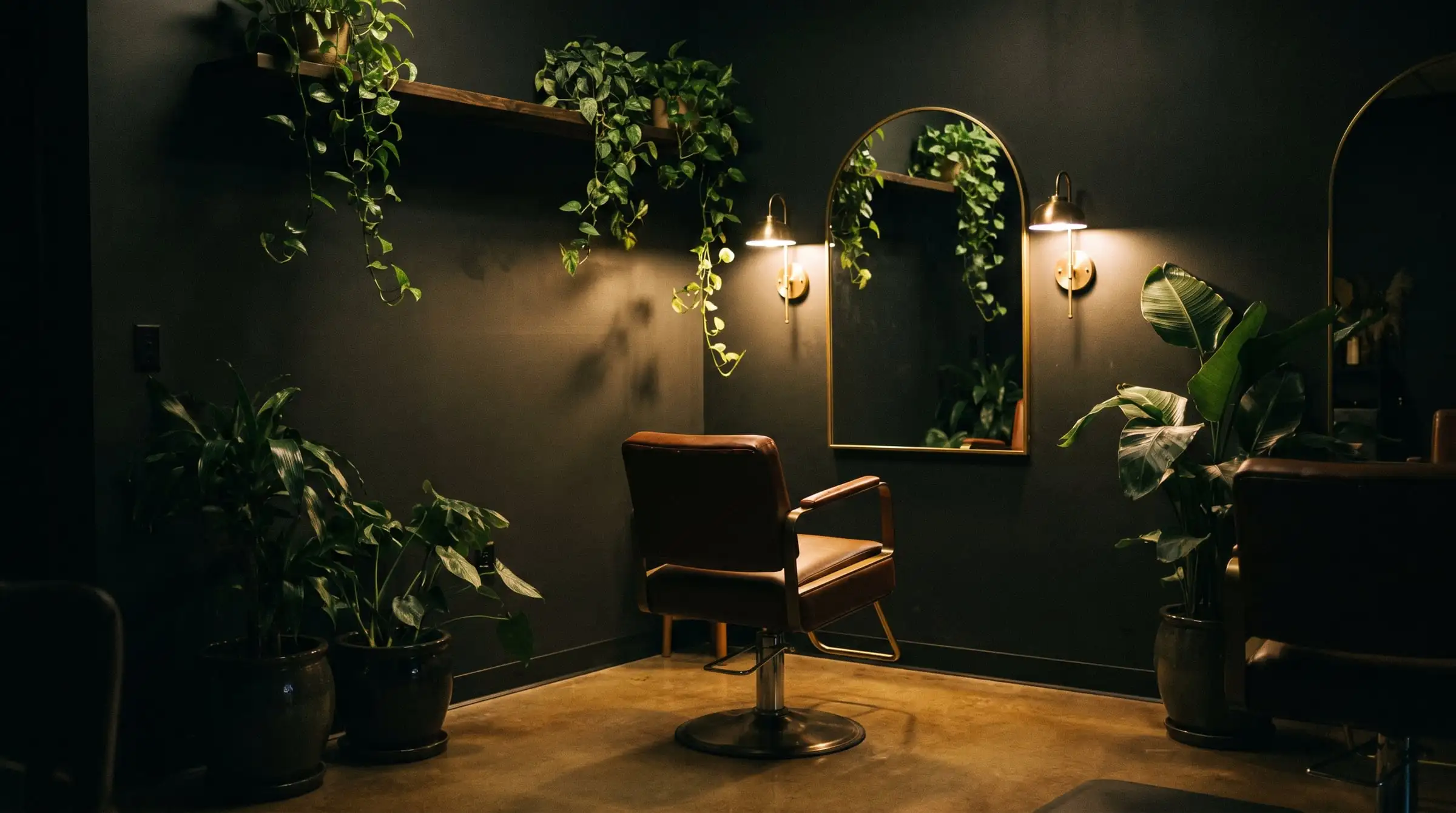 An intimate salon interior after dark with charcoal walls, an arched brass mirror, glowing brass wall lamps and green plants