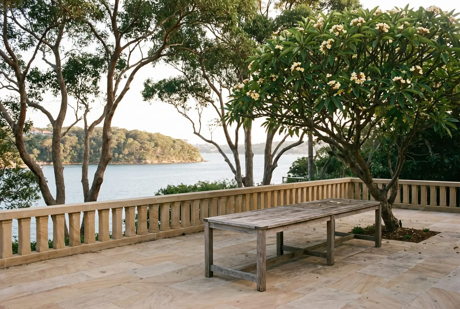 Sandstone terrace with a weathered timber table under a frangipani, overlooking the harbour