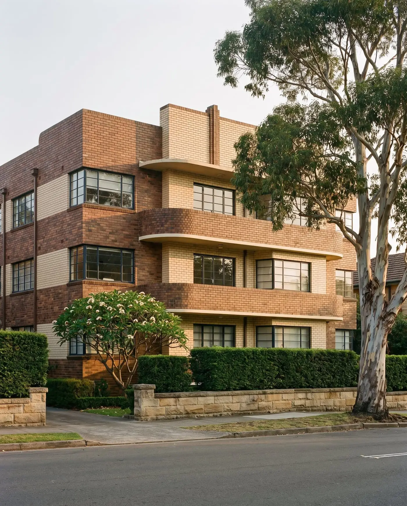 Three-storey Art Deco apartment building in two tones of brick with rounded balconies behind a sandstone wall