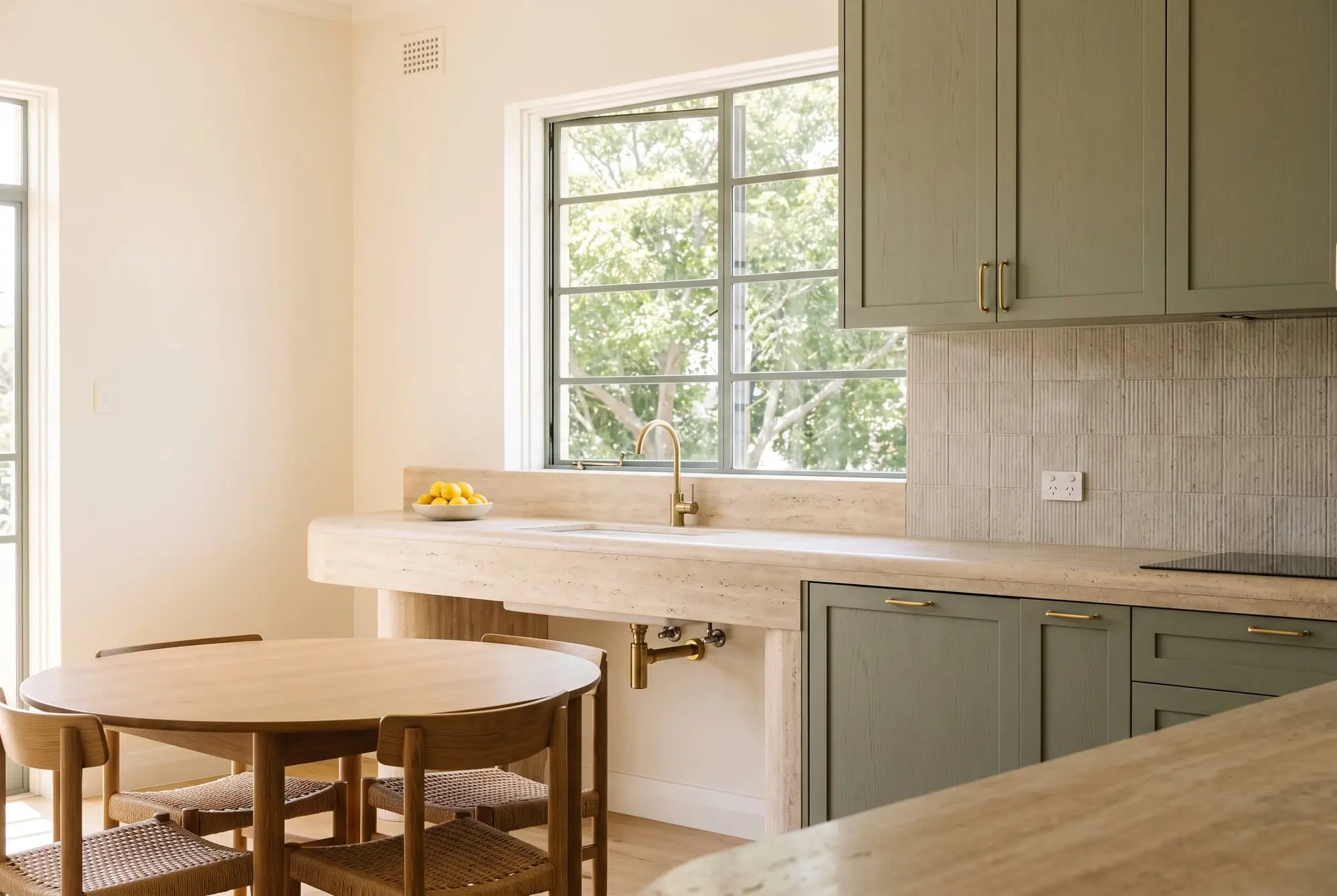 Kitchen with sage-green cabinetry, a travertine bench and a round oak dining table by a steel-framed window