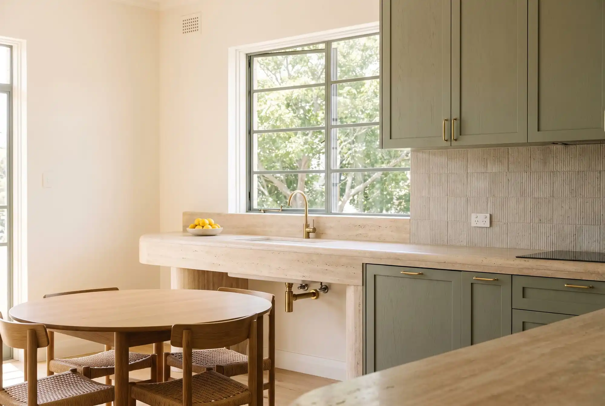 Kitchen with sage-green cabinetry, a travertine bench and a round oak dining table by a steel-framed window