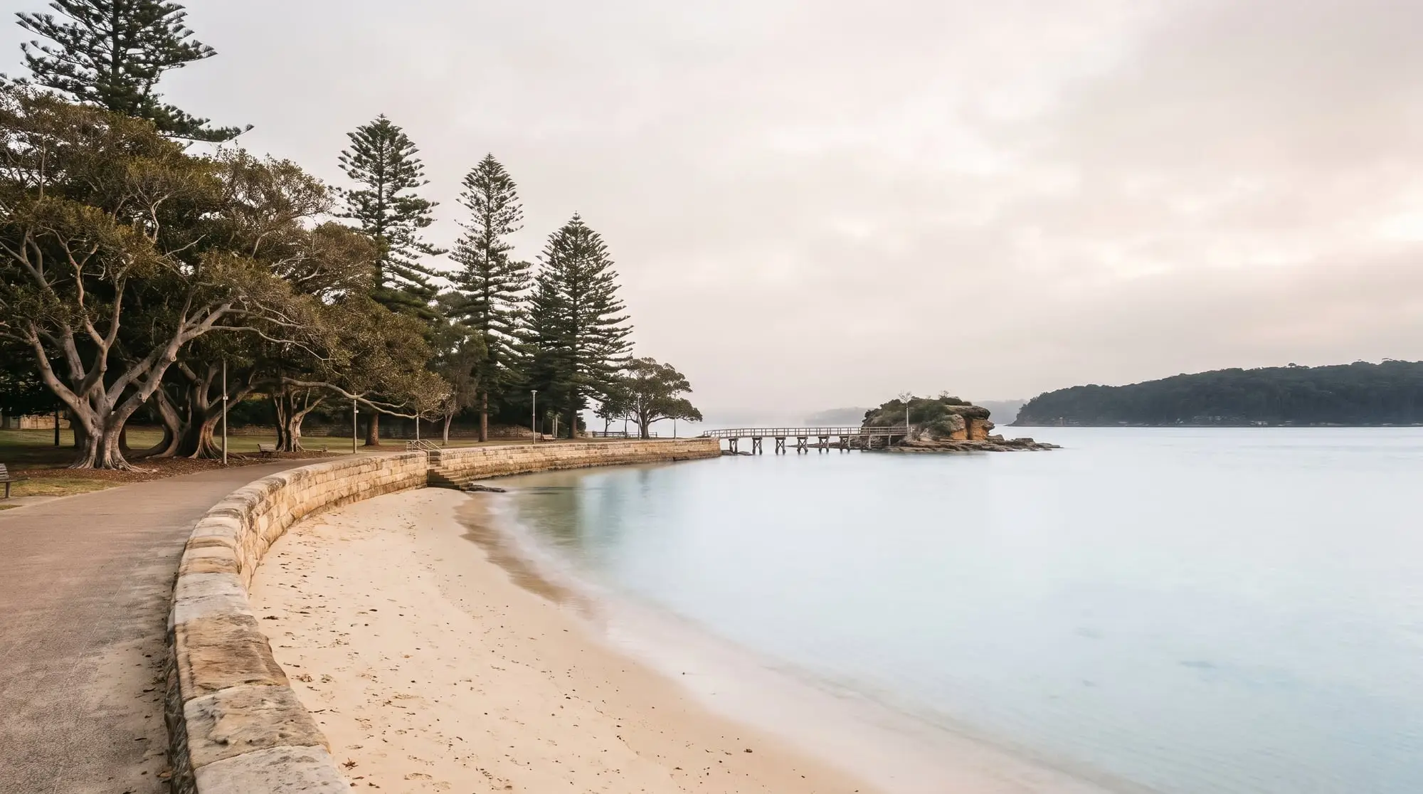 Calm harbour beach at dawn with a curved sandstone sea wall, fig trees and a footbridge to a small island