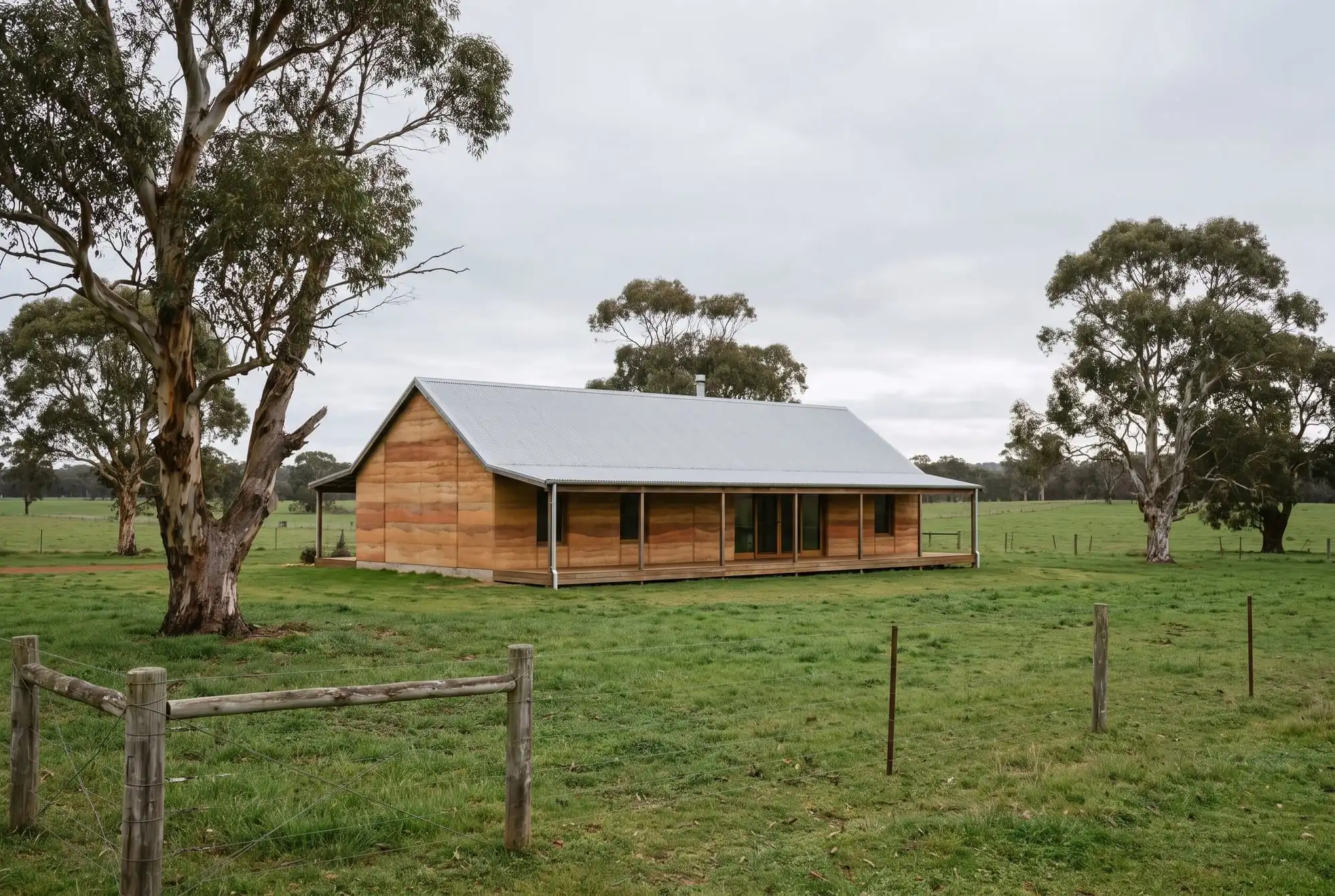 A gabled rammed earth farmhouse with a corrugated steel roof and wide verandah in a green paddock with old eucalypts