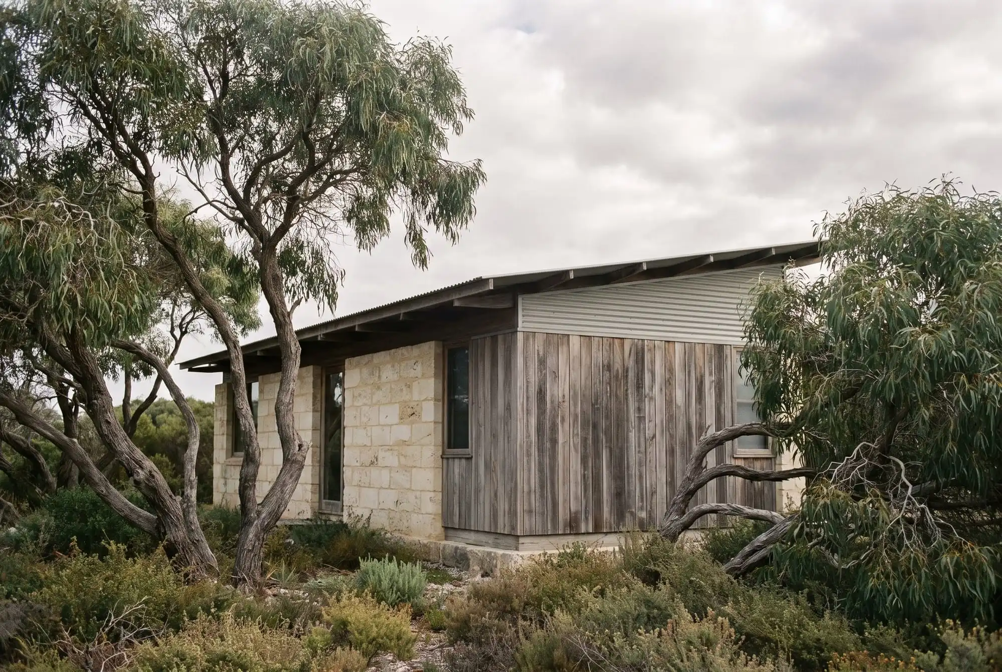 A low limestone and weathered timber house under gnarled peppermint trees with coastal heath in front