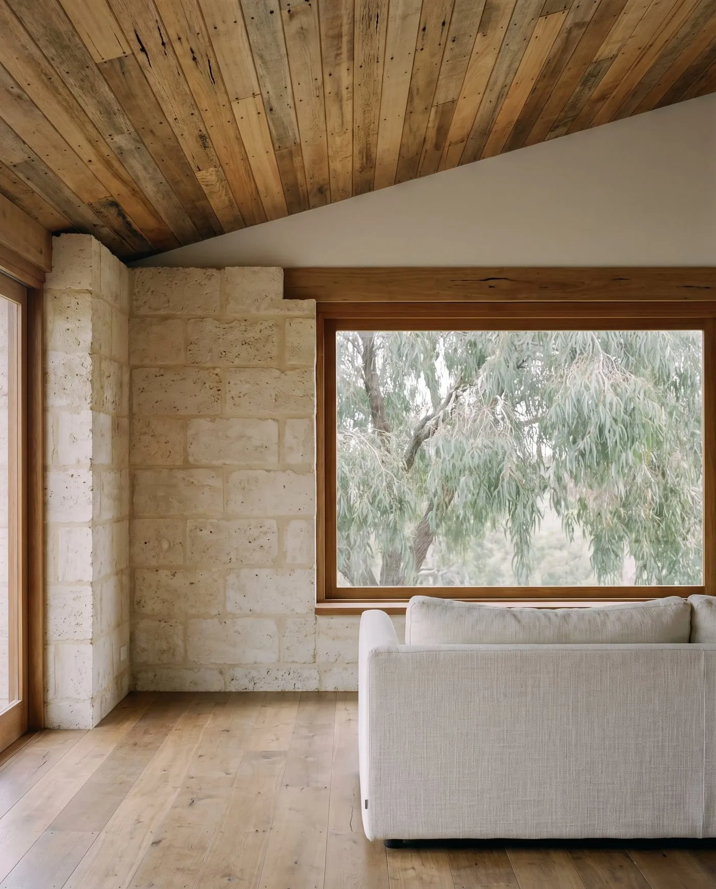 A living room with a recycled hardwood ceiling, a limestone block wall and a window framing peppermint tree leaves