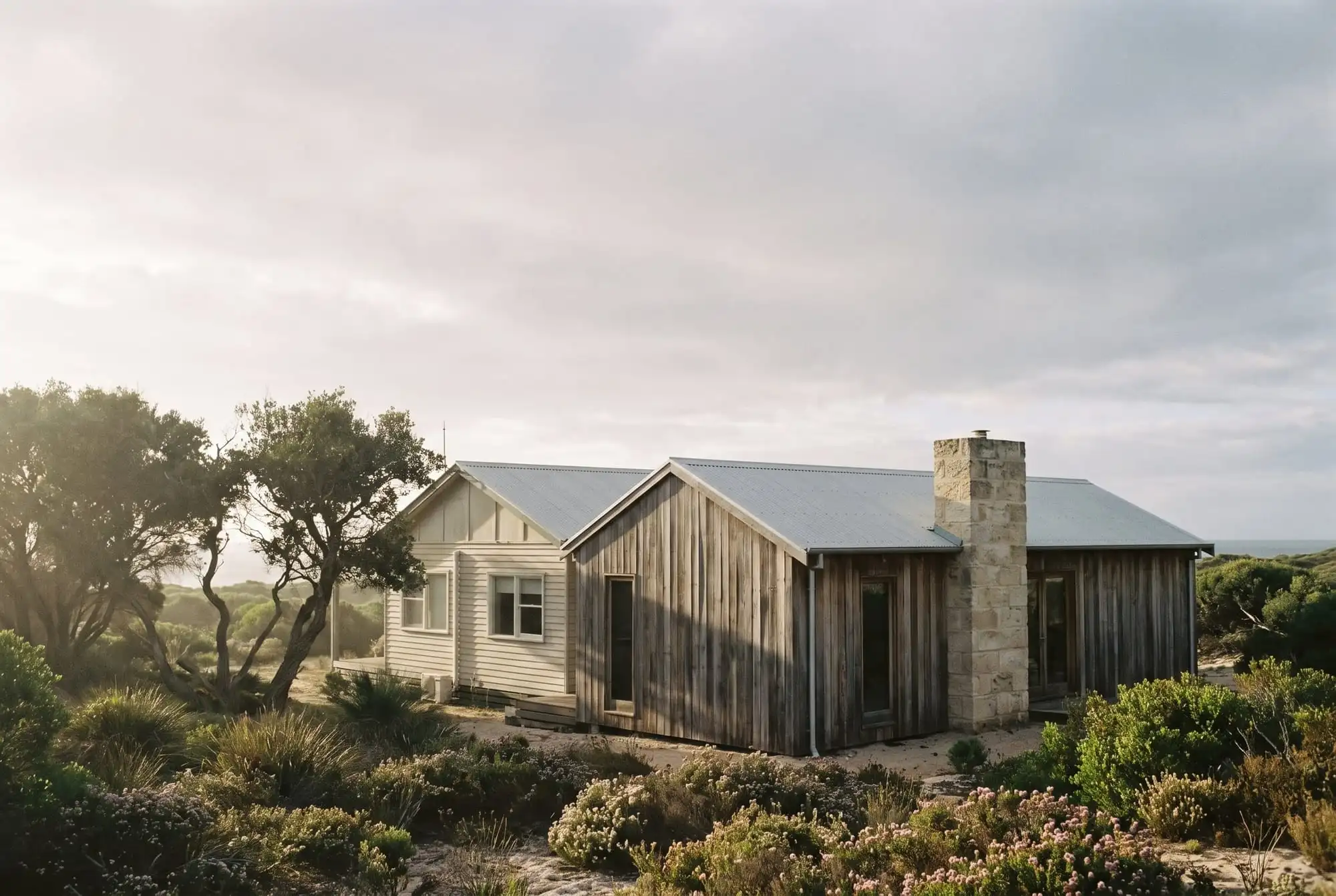 An old weatherboard beach shack beside a new wing clad in weathered recycled timber with a limestone chimney, in coastal heath