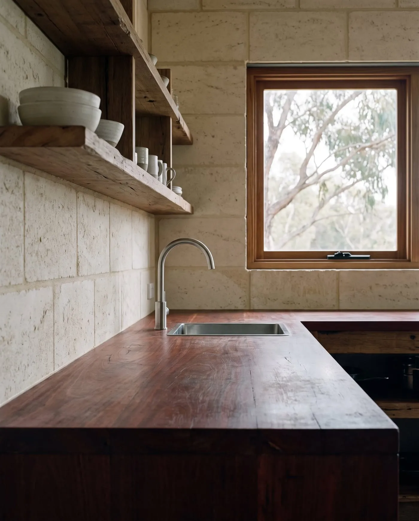A kitchen with a thick recycled jarrah benchtop, a limestone wall and a window to the eucalypts