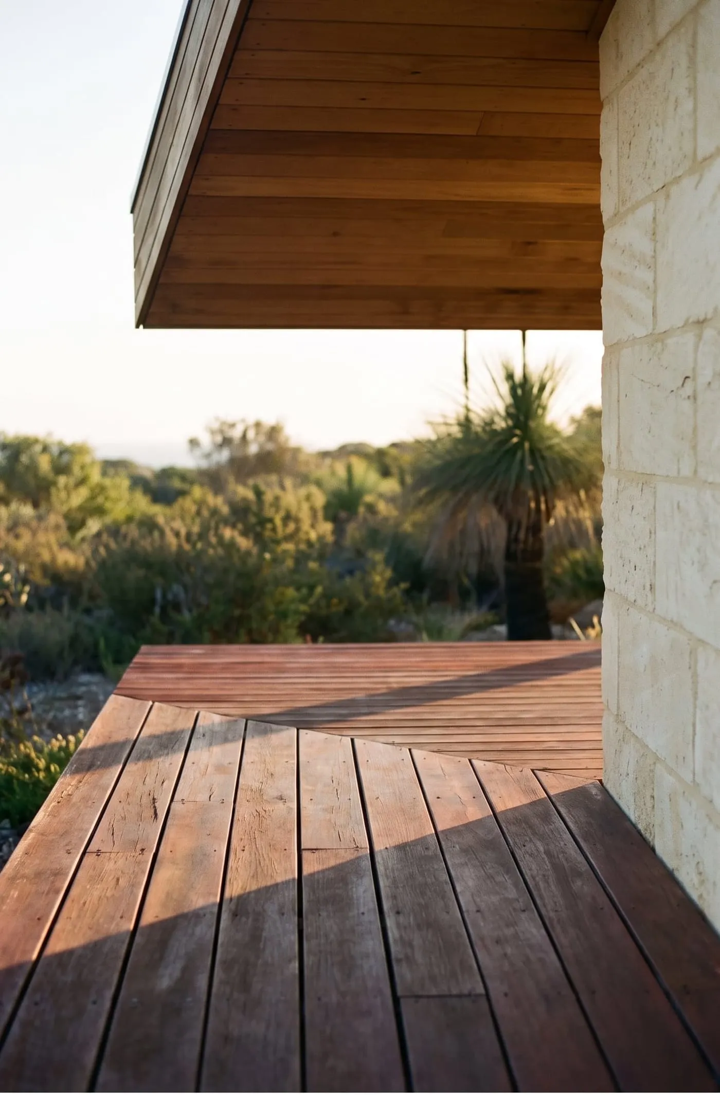A jarrah deck meeting a limestone wall under a timber-lined eave, with grass trees beyond in late sun