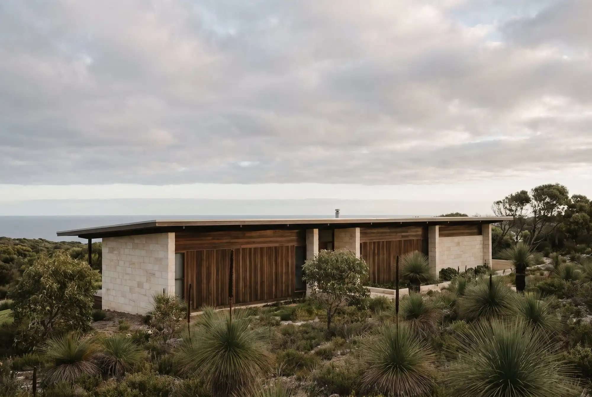 A long flat-roofed house of pale limestone and dark timber among grass trees on a coastal hillside, with the ocean behind