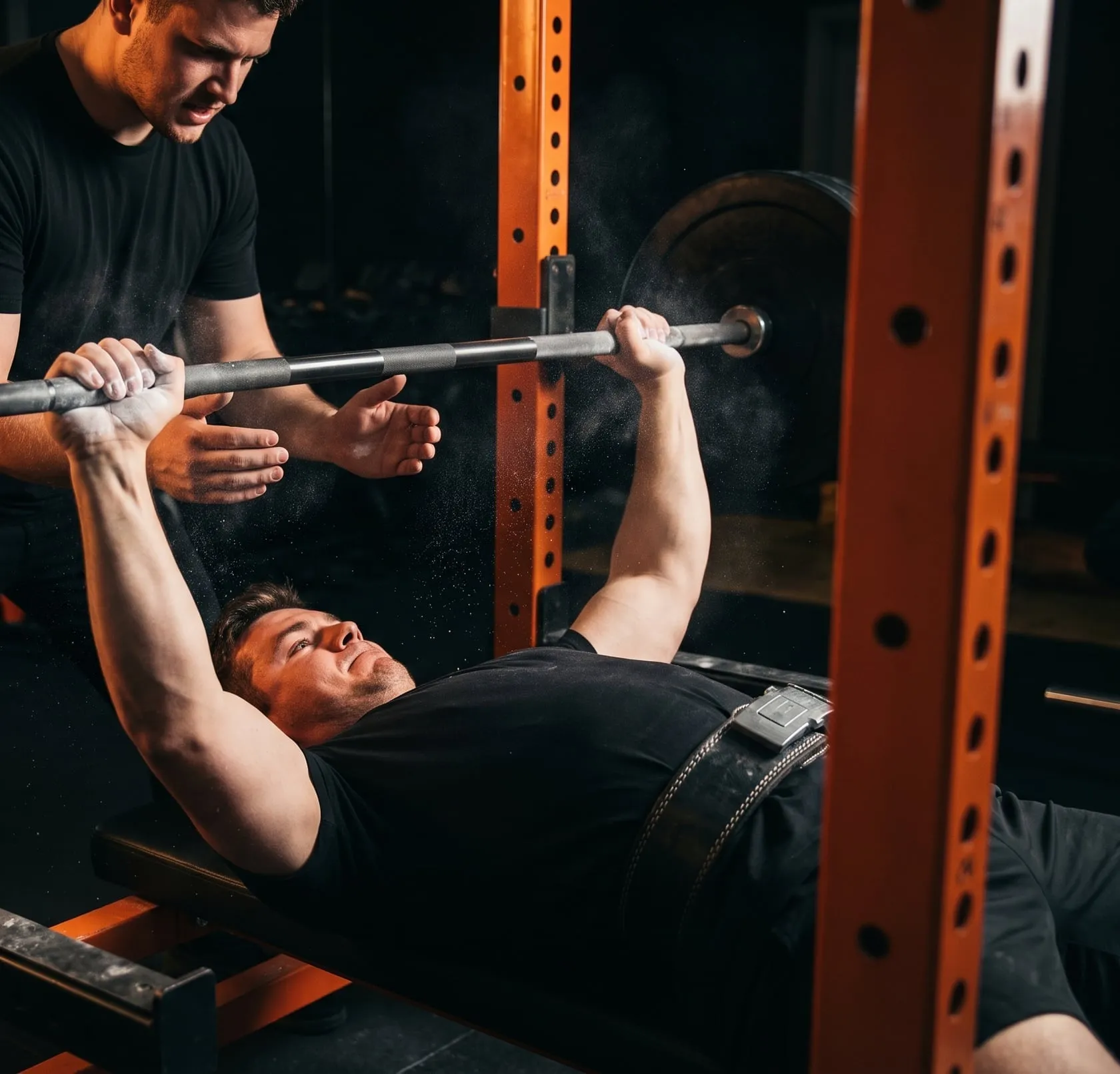 A lifter benching heavy in an orange rack while a spotter's hands hover under the bar