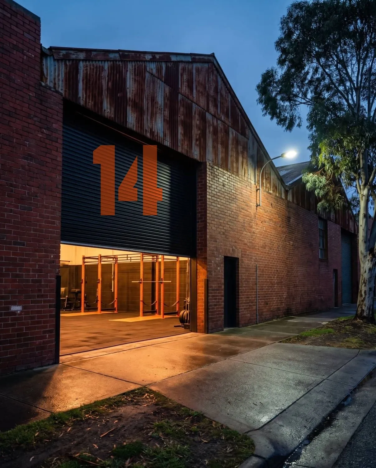 The warehouse before sunrise, its black roller door half raised under a giant orange painted 14, warm light spilling out