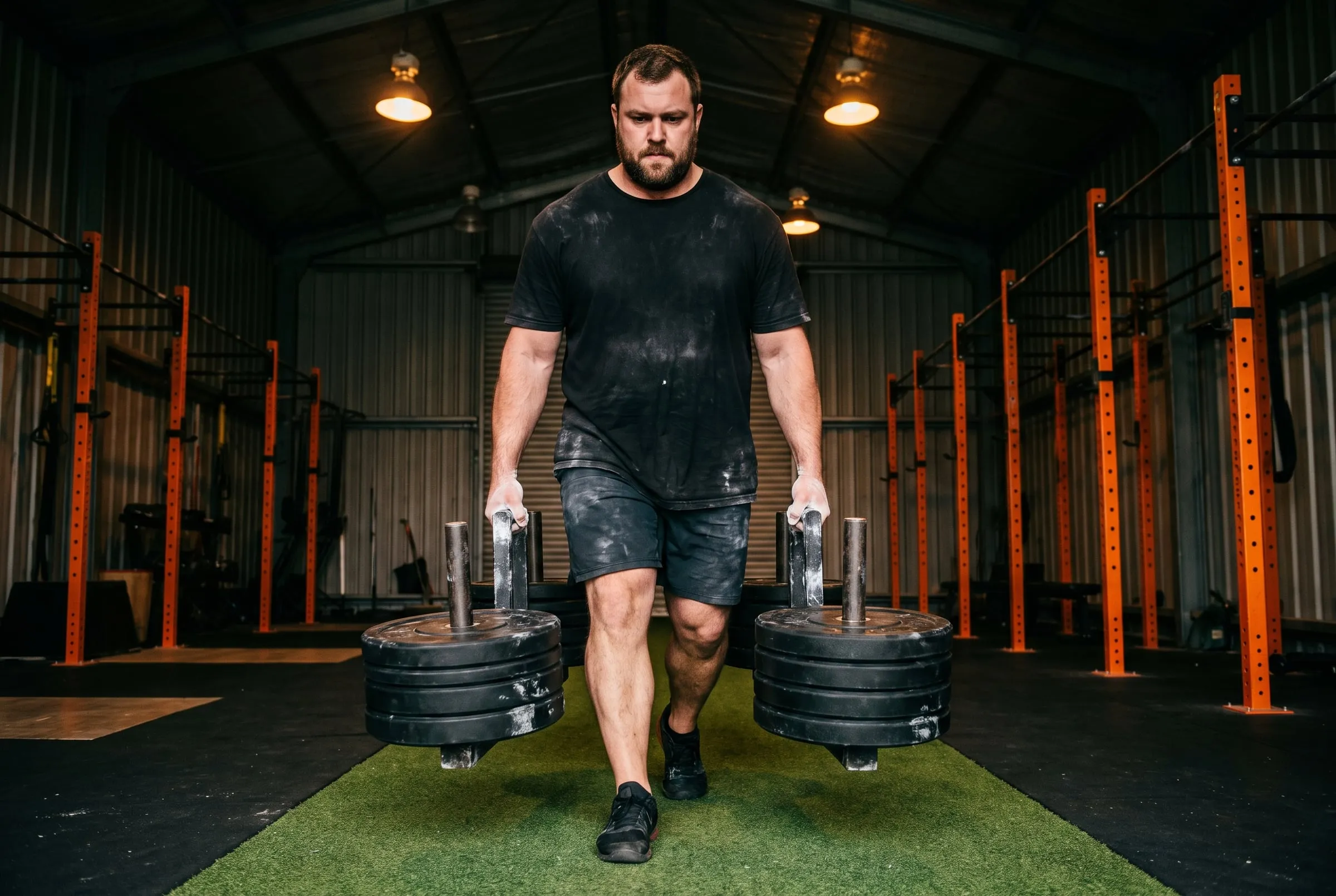 A big man walking a heavy farmer's carry down a strip of turf between orange racks
