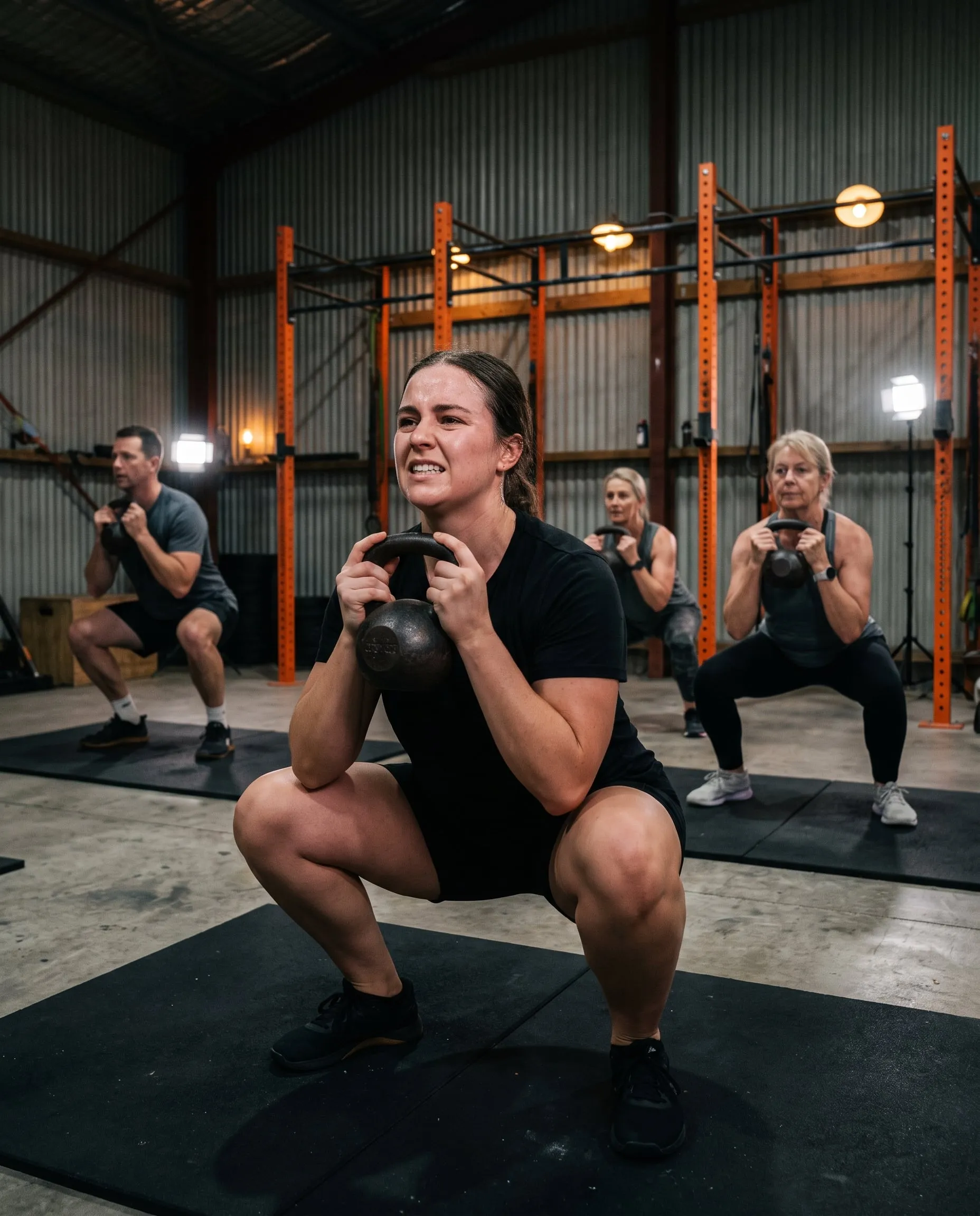 A woman deep in a kettlebell goblet squat with a small class doing the same behind her