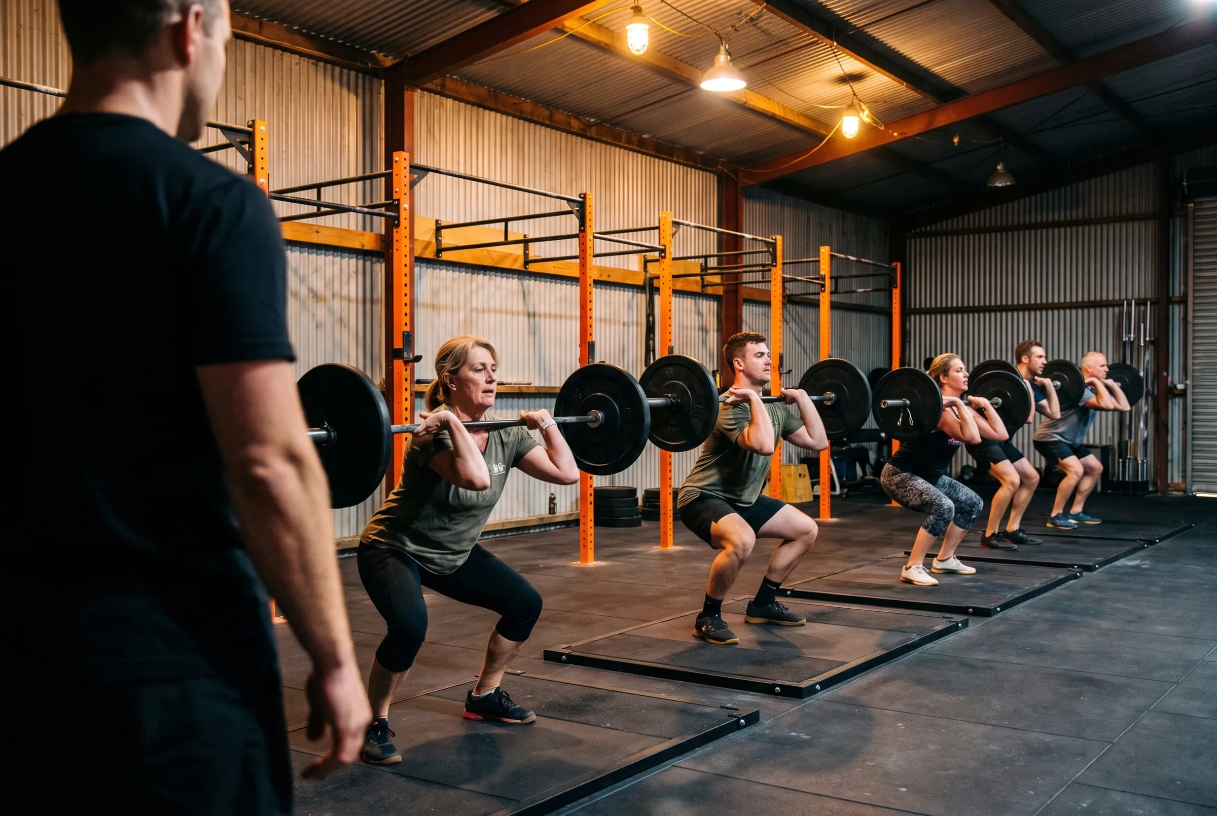 A small class front squatting in a row on black platforms while a coach watches from the side