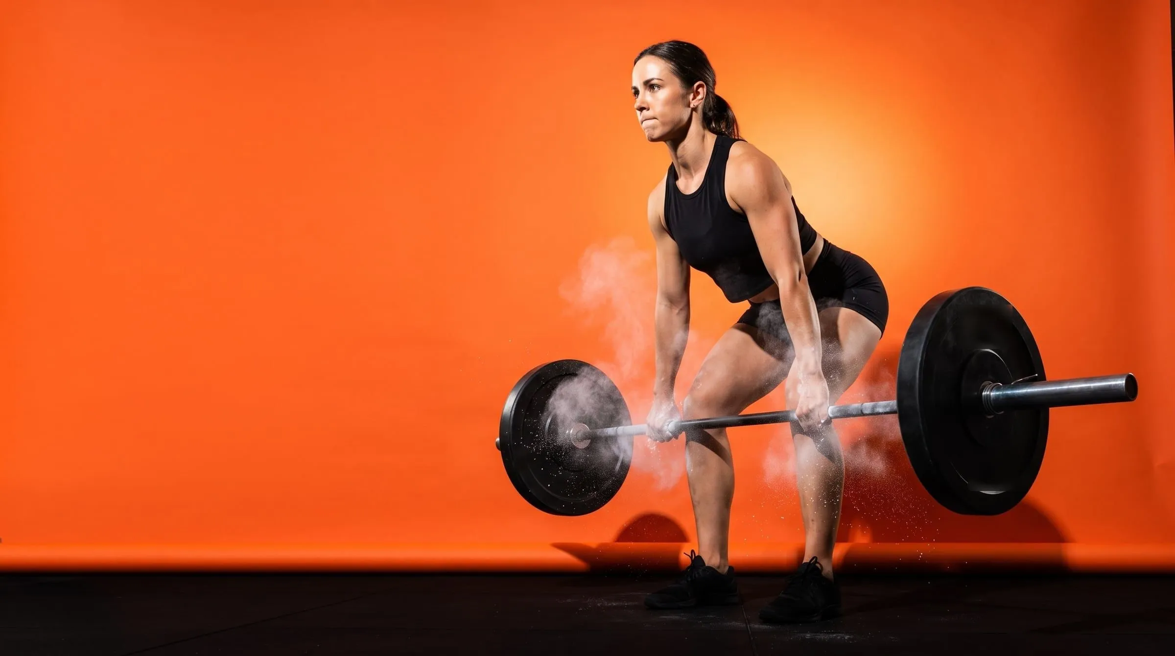 A woman mid-deadlift against an orange backdrop, chalk bursting off her hands as the bar passes her knees