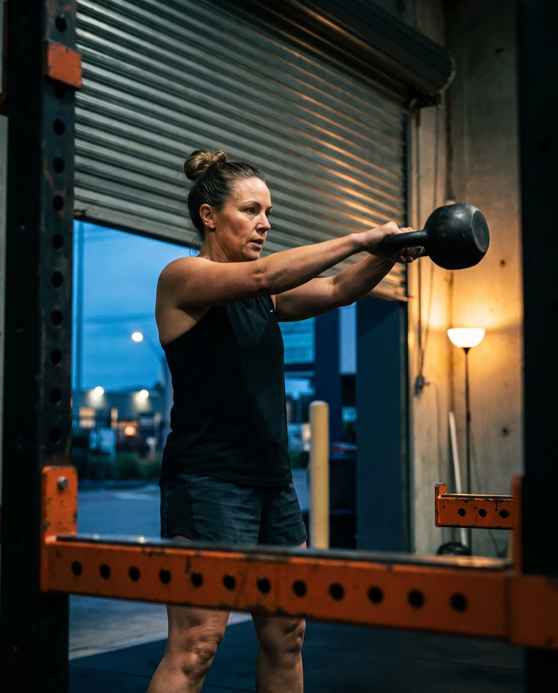 A woman in her forties swinging a kettlebell at dawn in front of a half-open roller door