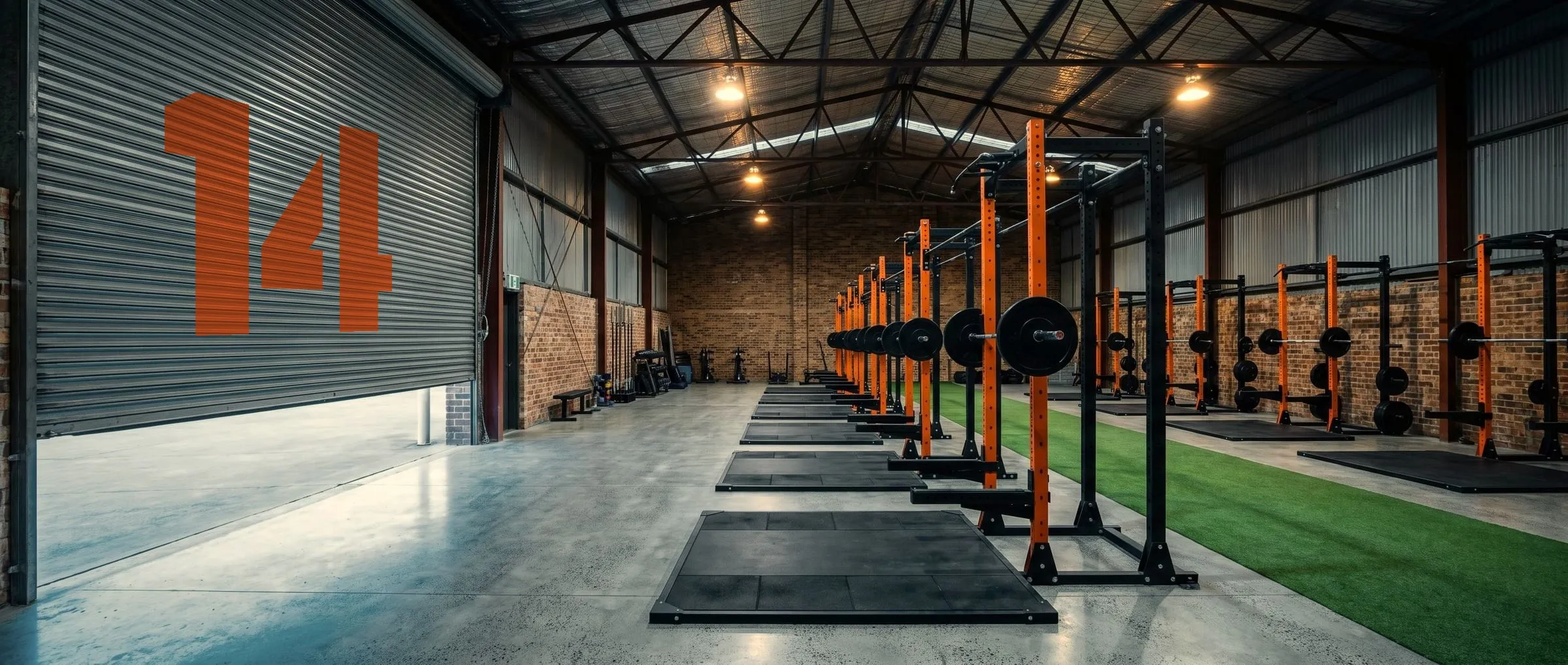 An empty warehouse gym at dawn, a long row of orange squat racks beside green turf, and a giant orange 14 painted on the half-raised roller door