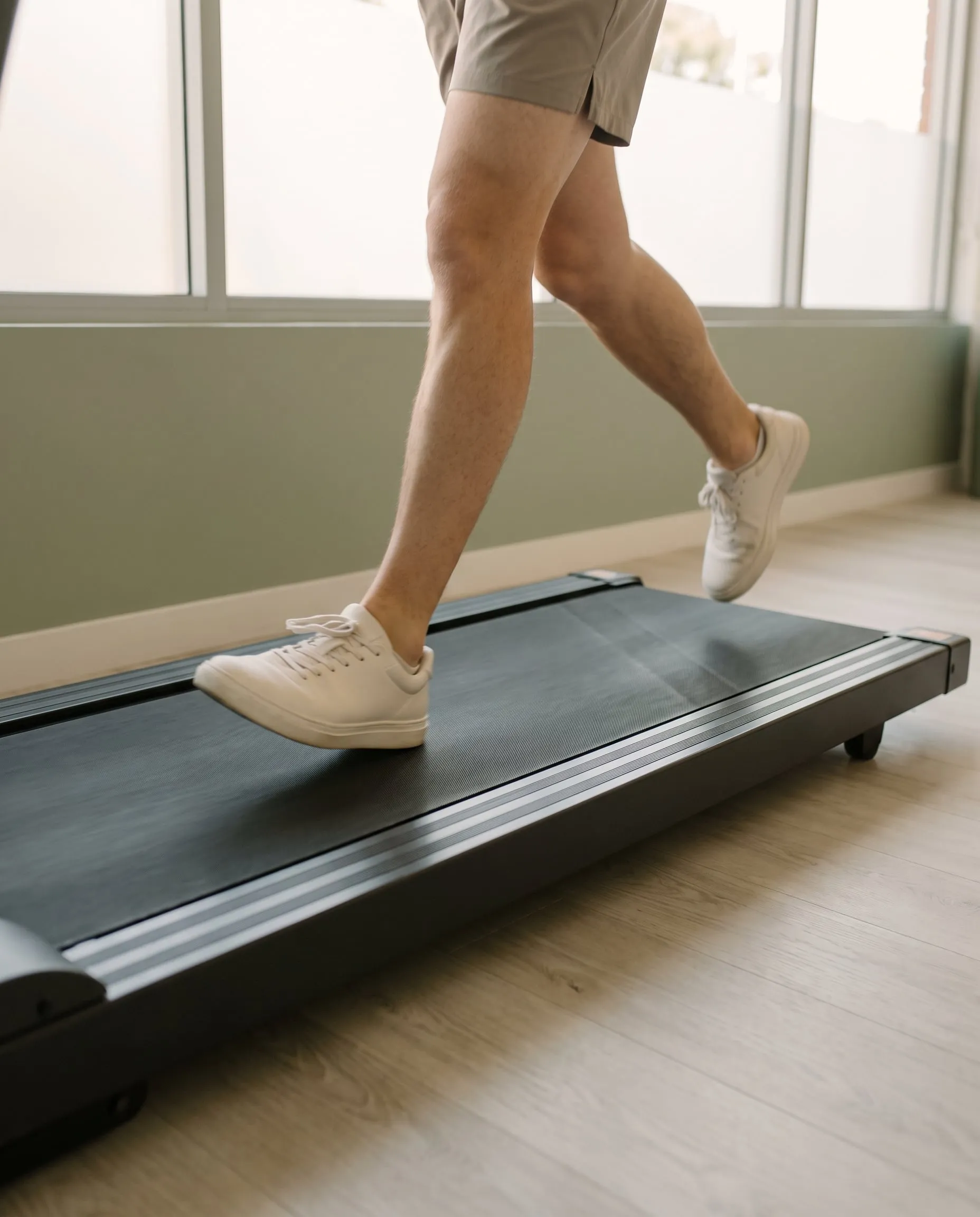 A runner's lower legs and white shoes mid-stride on a treadmill in a sage green studio
