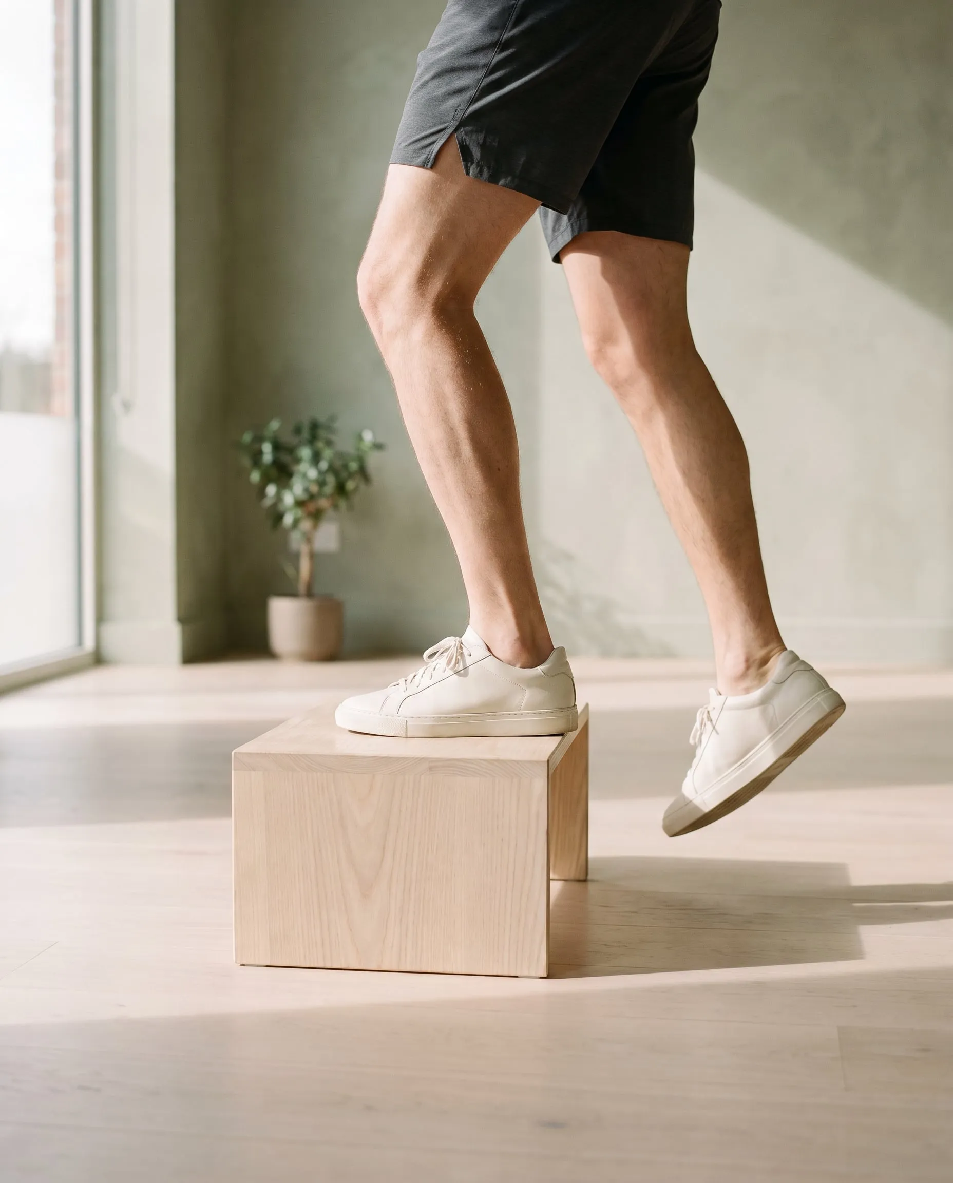 Legs in dark shorts stepping up onto a pale timber box in a sunlit studio
