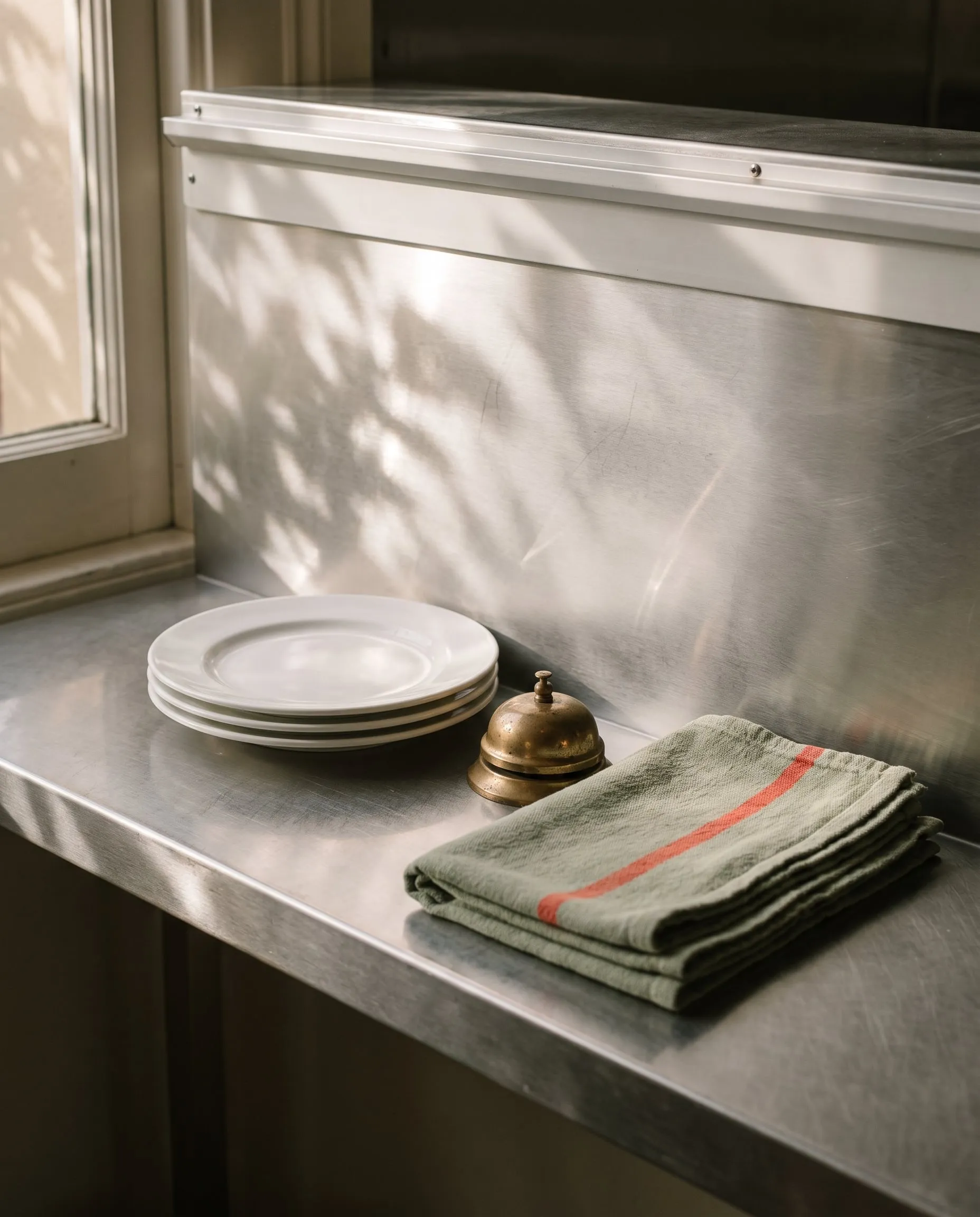 Stacked white plates, a brass service bell and a folded tea towel on a stainless steel kitchen pass