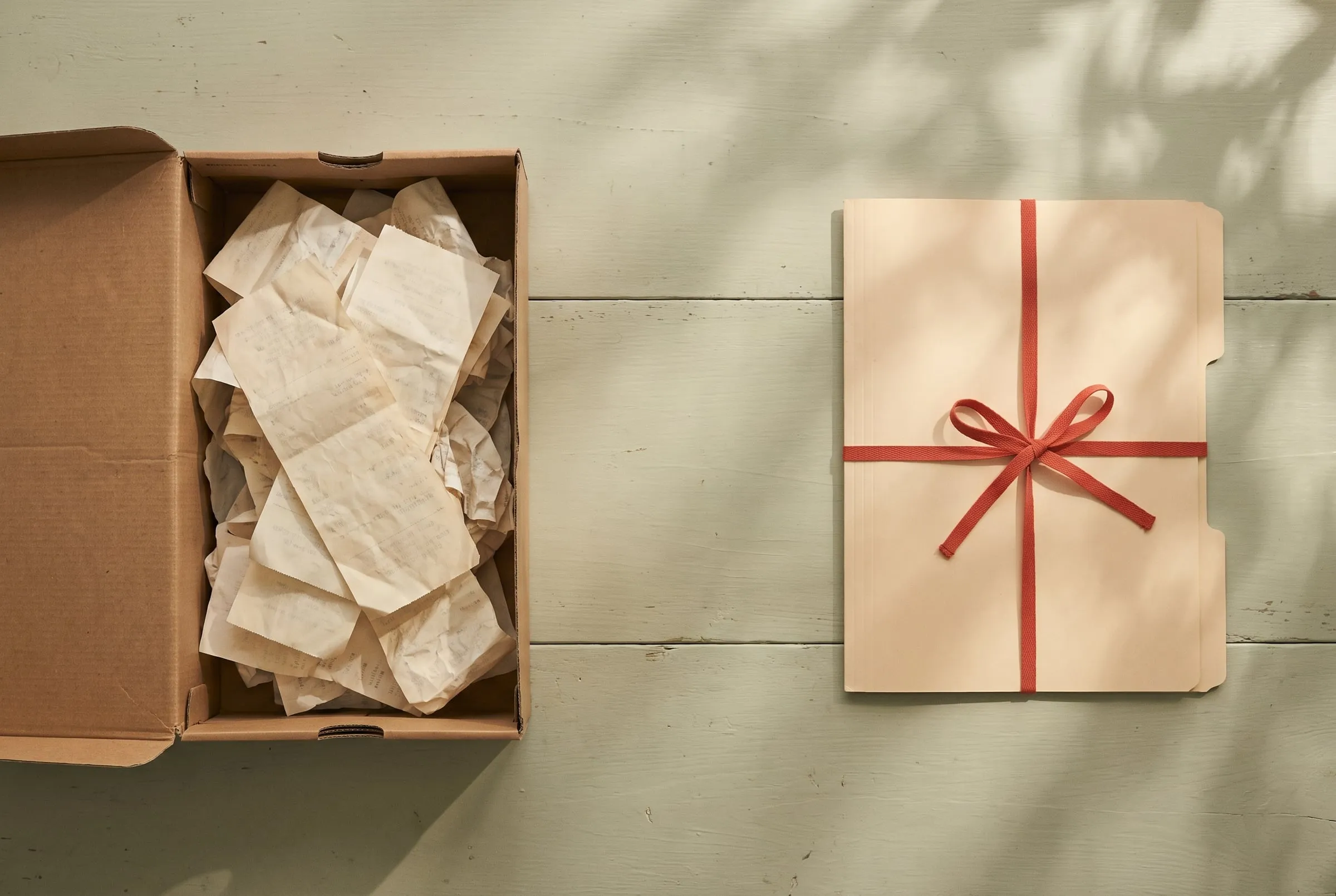 A shoebox overflowing with faded receipts next to a neat manila folder tied with red tape