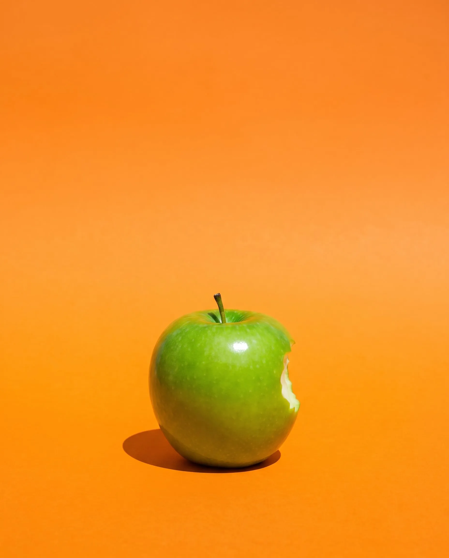 A green apple with one small bite taken out of it, on a bright tangerine background