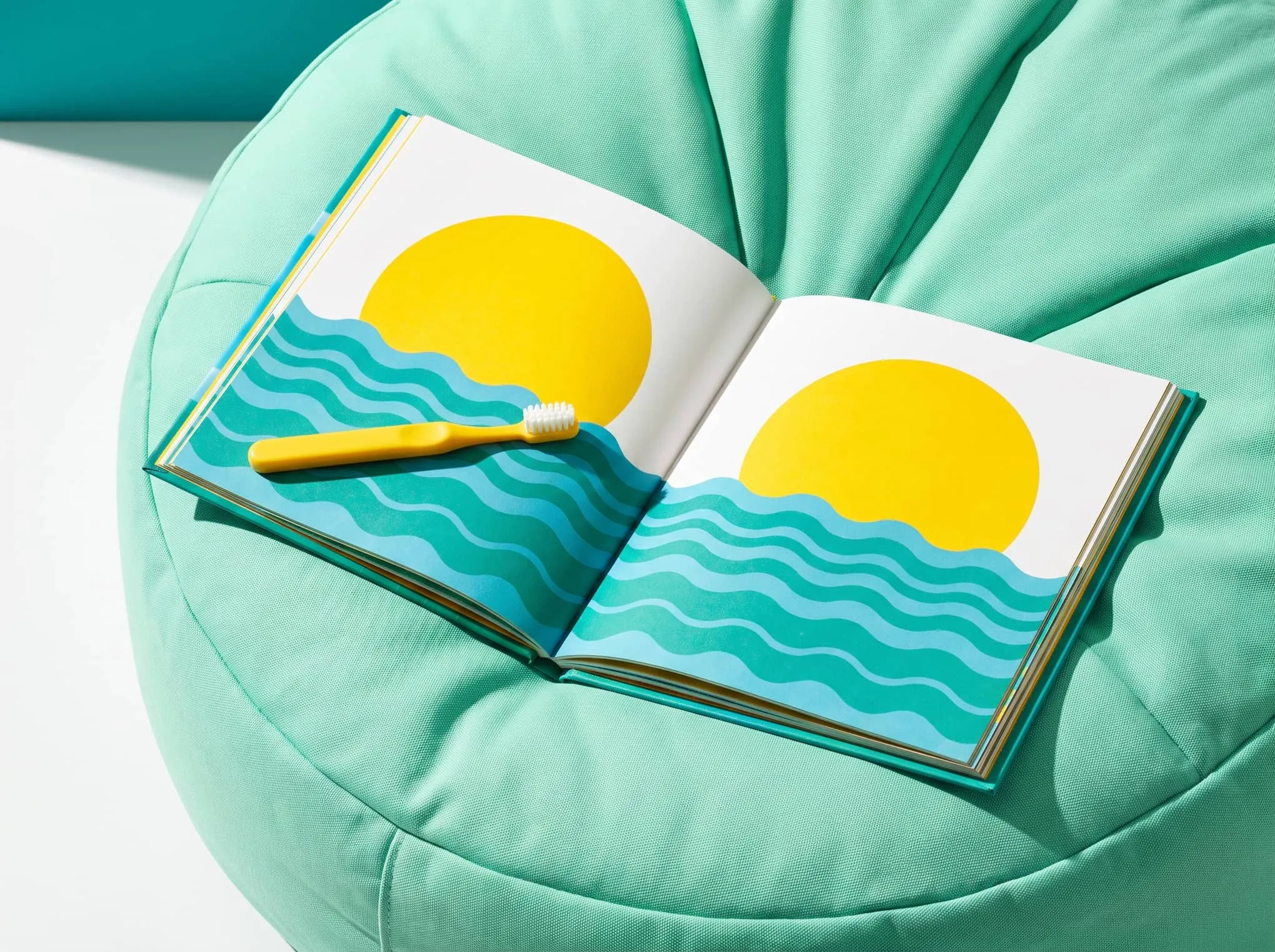 An open picture book of a yellow sun over blue waves on a mint beanbag, with a yellow toothbrush resting on it