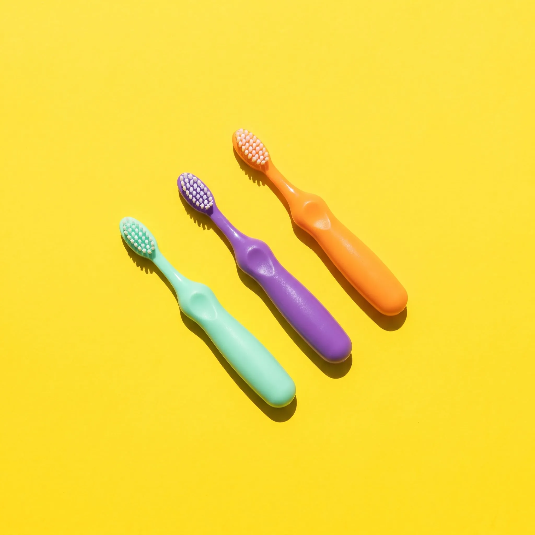 Three small toothbrushes in mint, grape and tangerine on sunny yellow paper