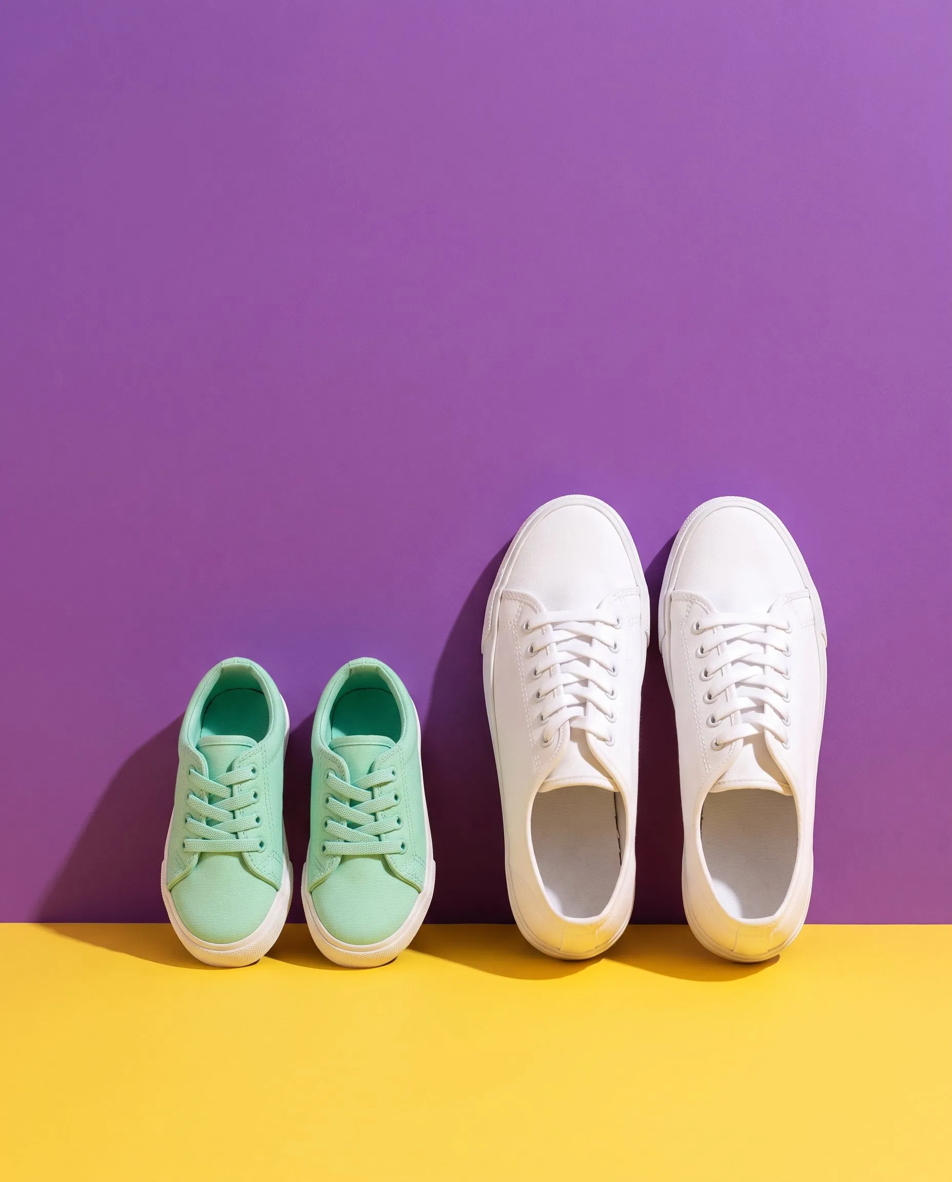 A pair of small mint sneakers beside a pair of grown-up white sneakers on a yellow floor