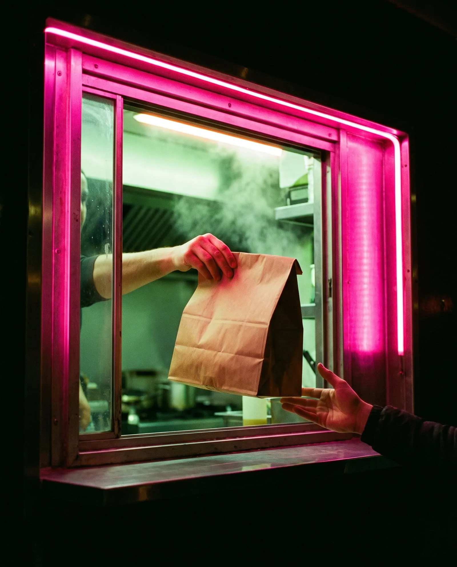 A paper takeaway bag handed out through a steel hatch lit with pink neon