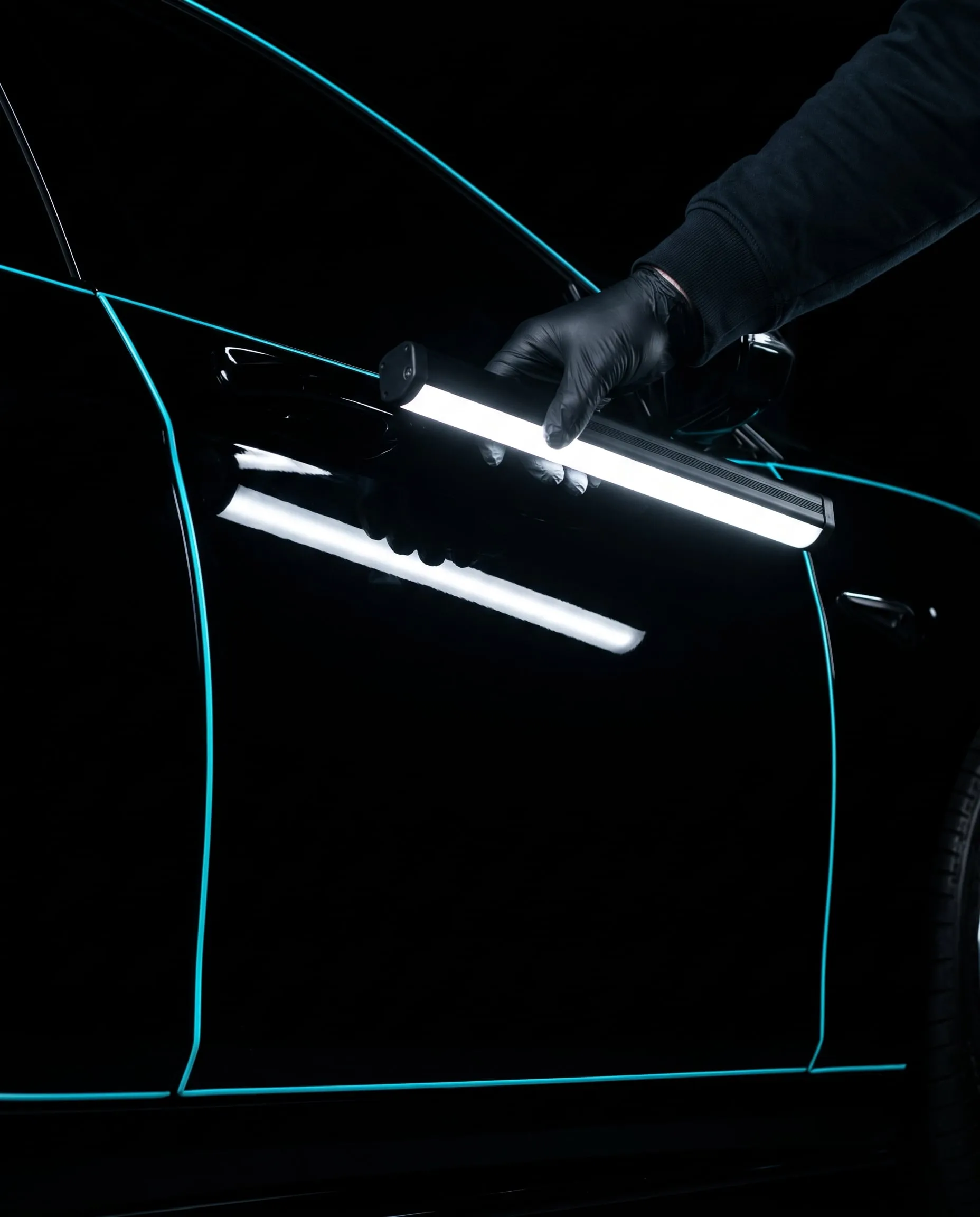 A gloved hand holding a linear LED inspection lamp against a gloss black door, its light reflected as one crisp line
