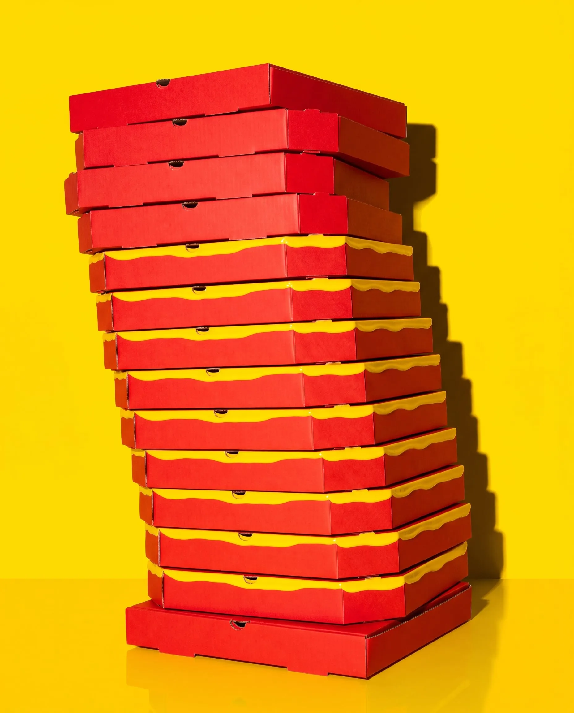 A tall leaning stack of red pizza boxes with a yellow stripe on a yellow background