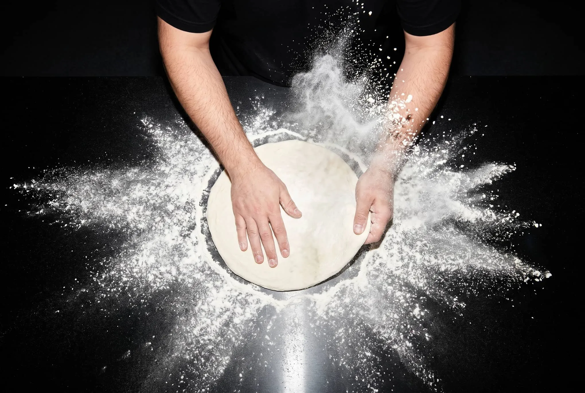 Hands stretching a round of pizza dough in a burst of flour on a black bench