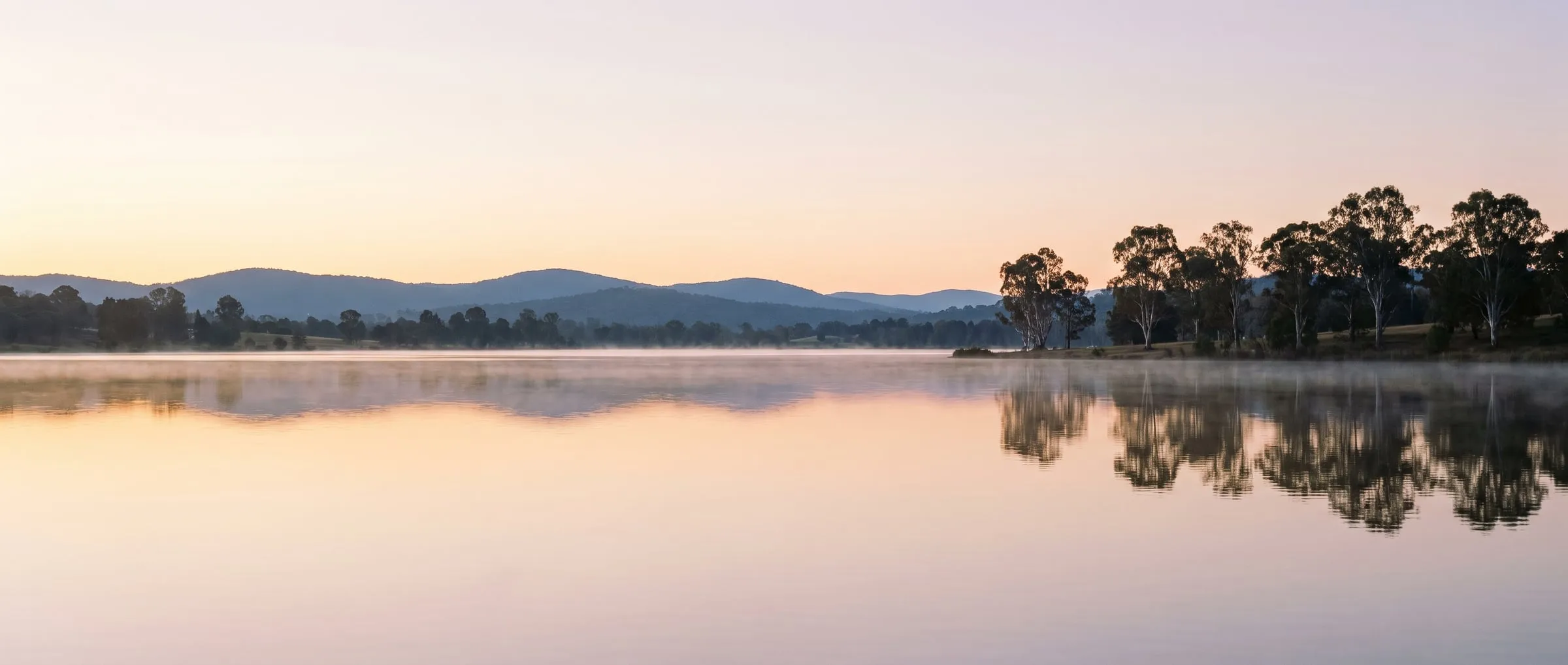 A still lake at first light, with gum trees on the far shore and soft hills beyond.