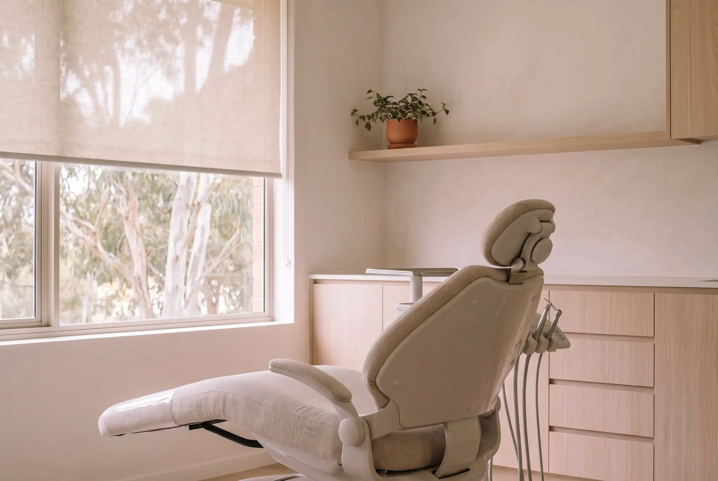 A calm treatment room with an oat upholstered chair, oak cabinetry and a window onto gum trees.