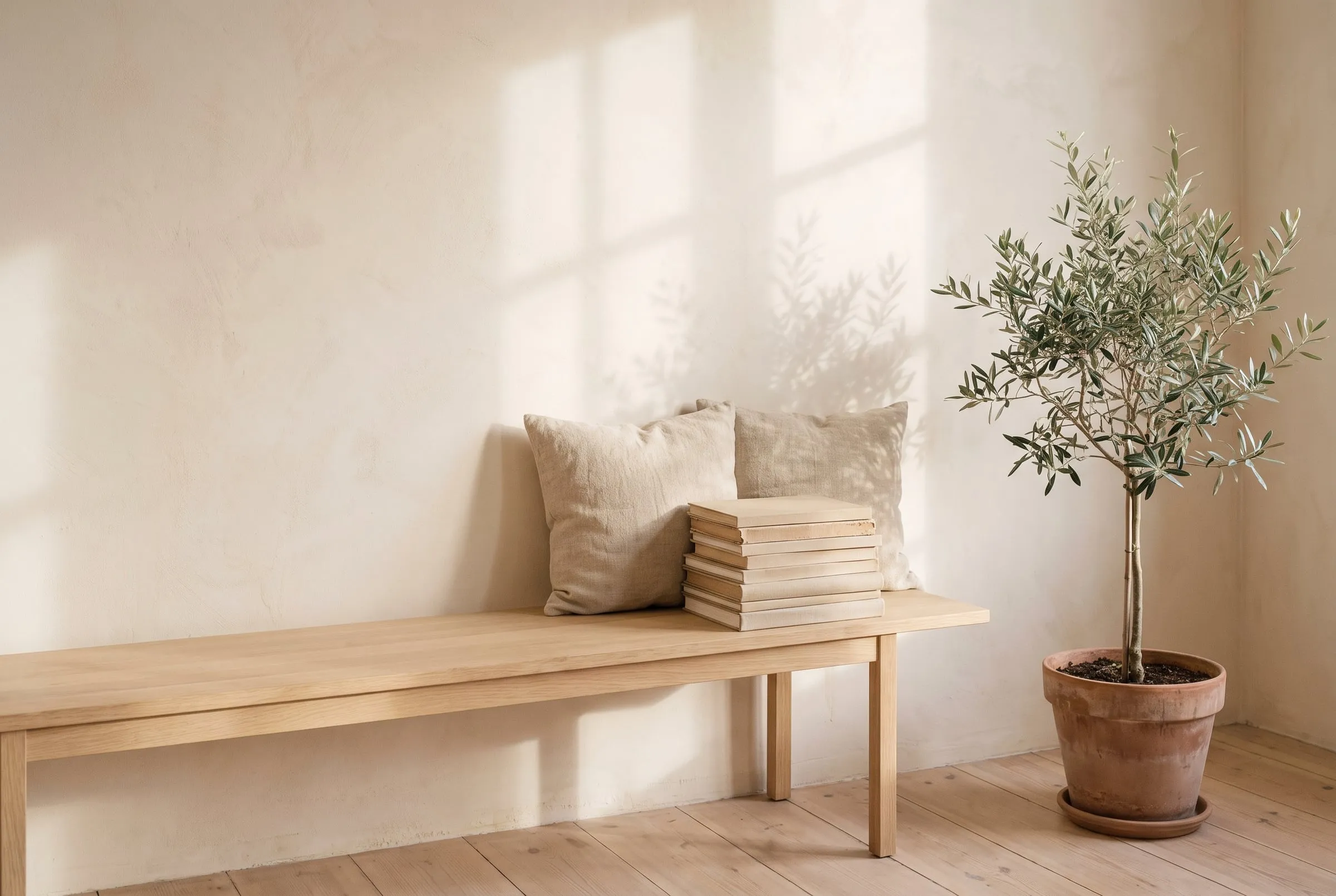An oak bench with linen cushions and a stack of books beside a potted olive tree.