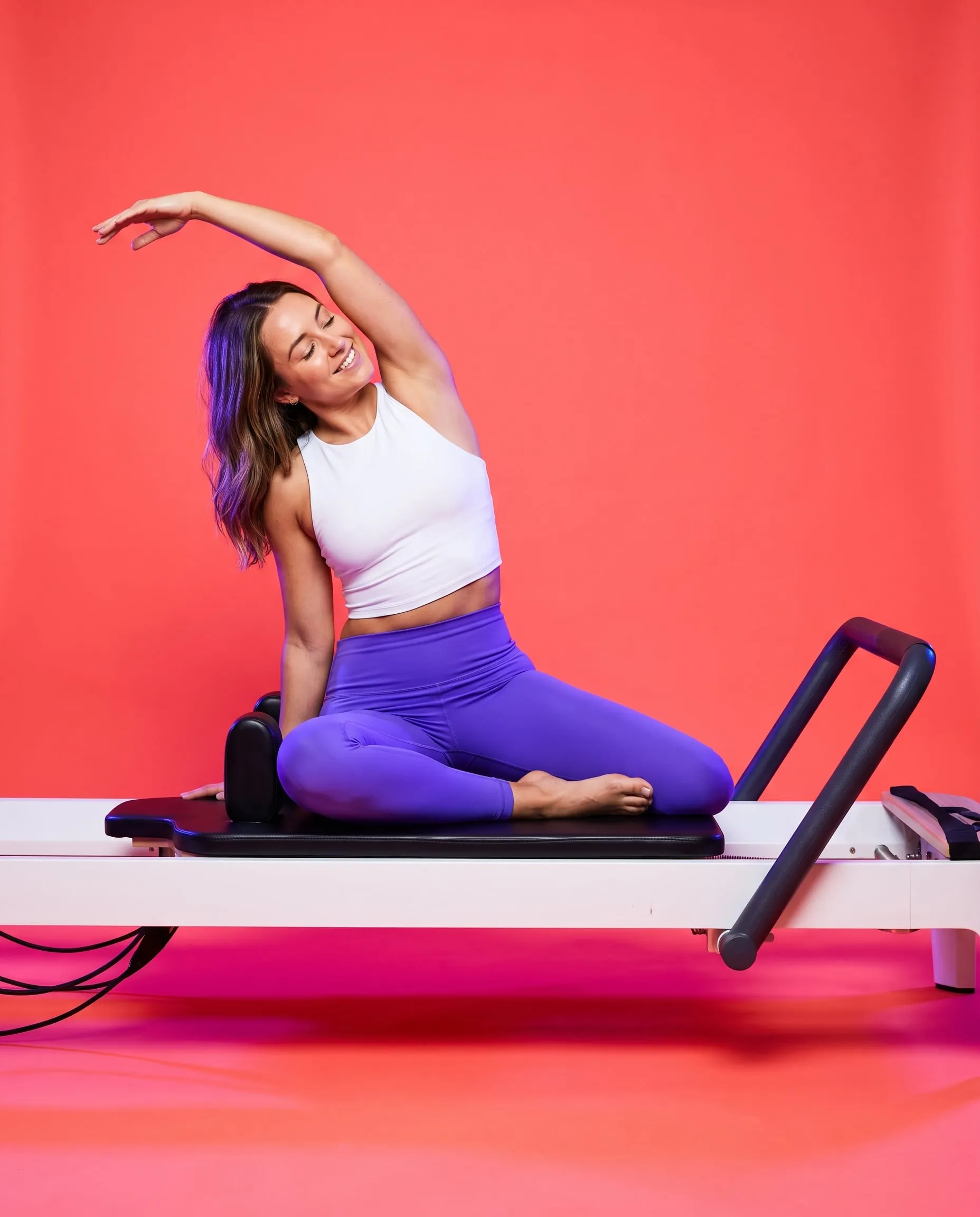 A woman in a side stretch on a reformer carriage, smiling with her eyes closed