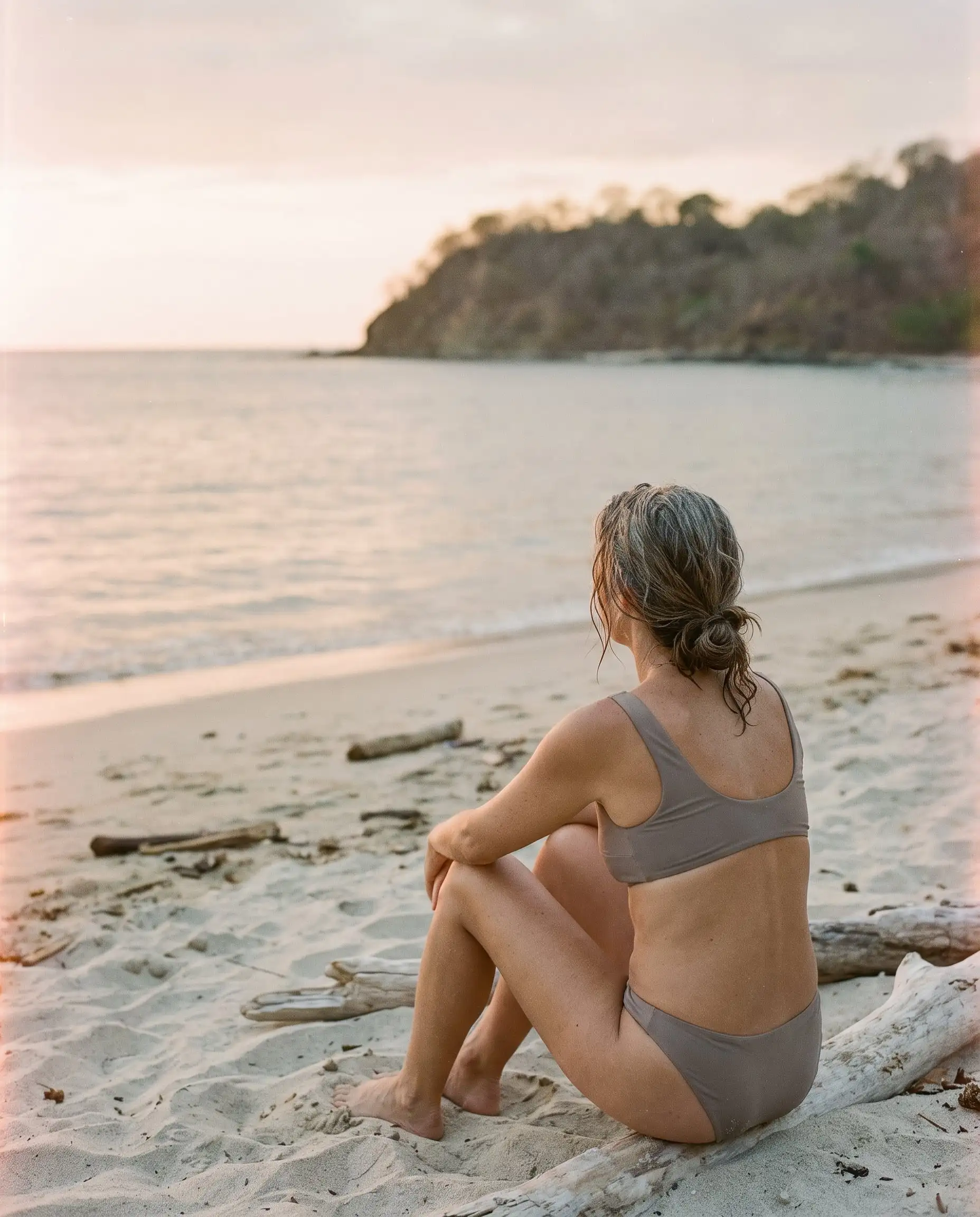 A swimmer in a Driftwood bikini sitting on driftwood at the edge of a beach, looking out at a calm sea
