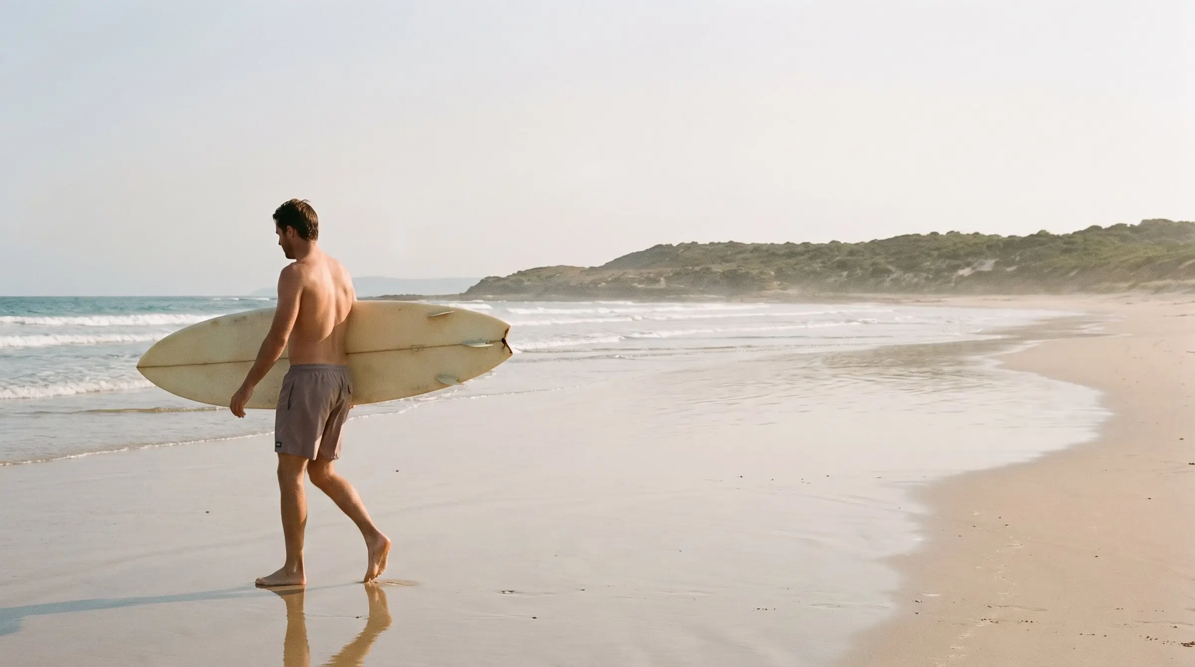 A surfer in Driftwood trunks carrying a cream single fin across firm sand towards the water